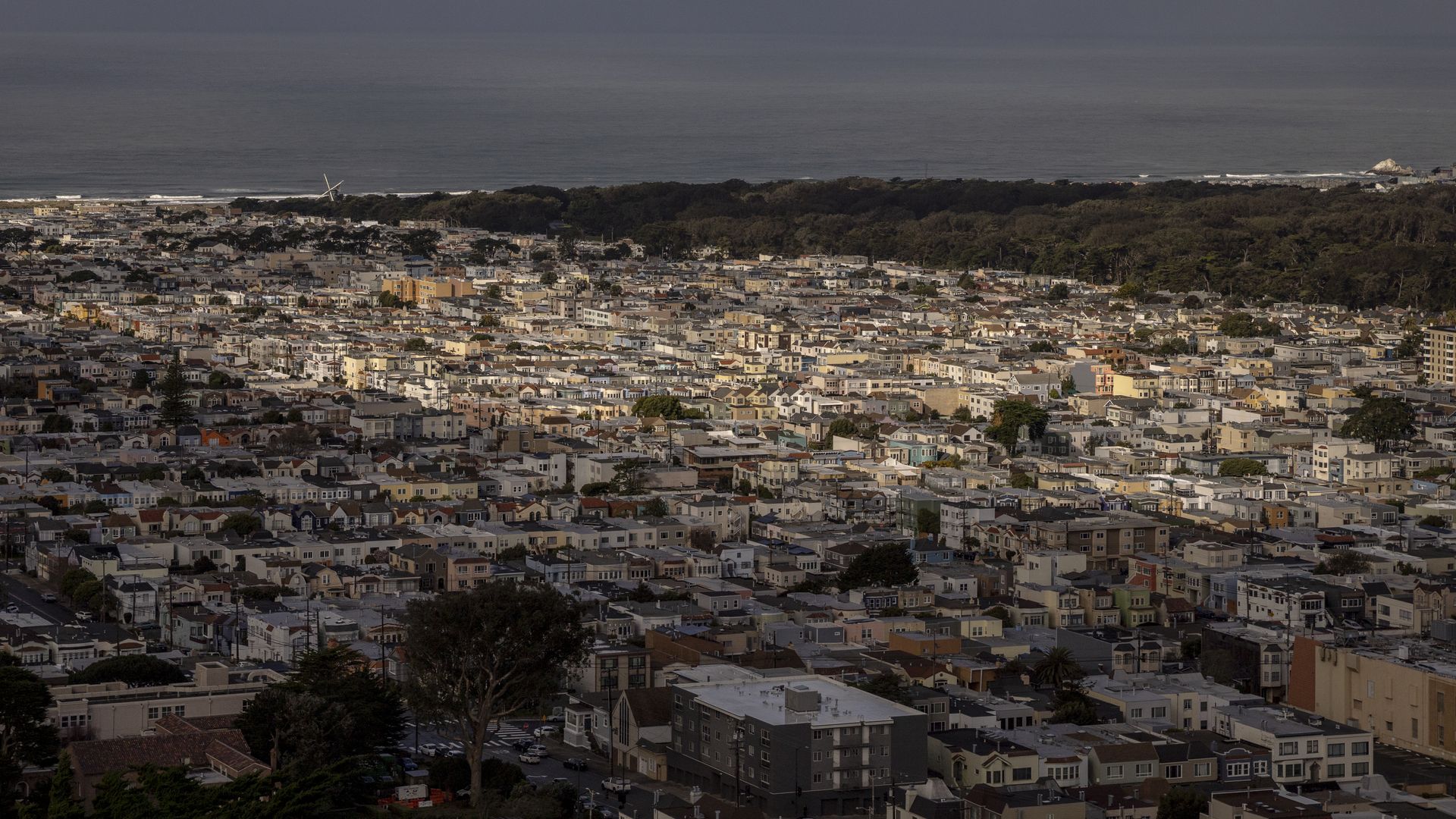 A densely packed cityscape with many houses under a cloudy sky, a forested area, and the ocean in the background with calm waves.