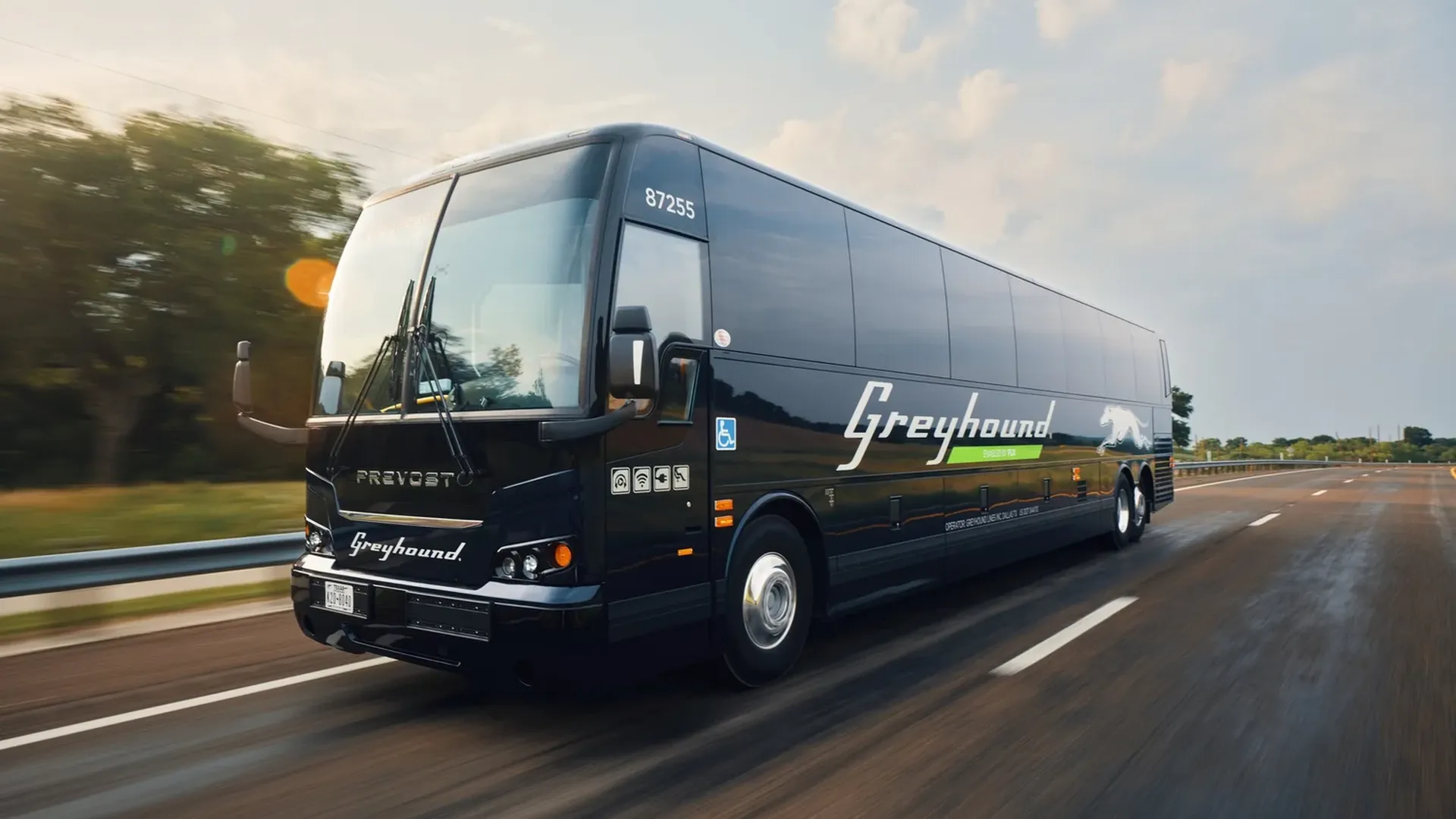 Black Greyhound bus speeding on a highway, white Greyhound logo and dog emblem along the side, Prevost badge on the front. Motion blur with a sun flare and blue sky.