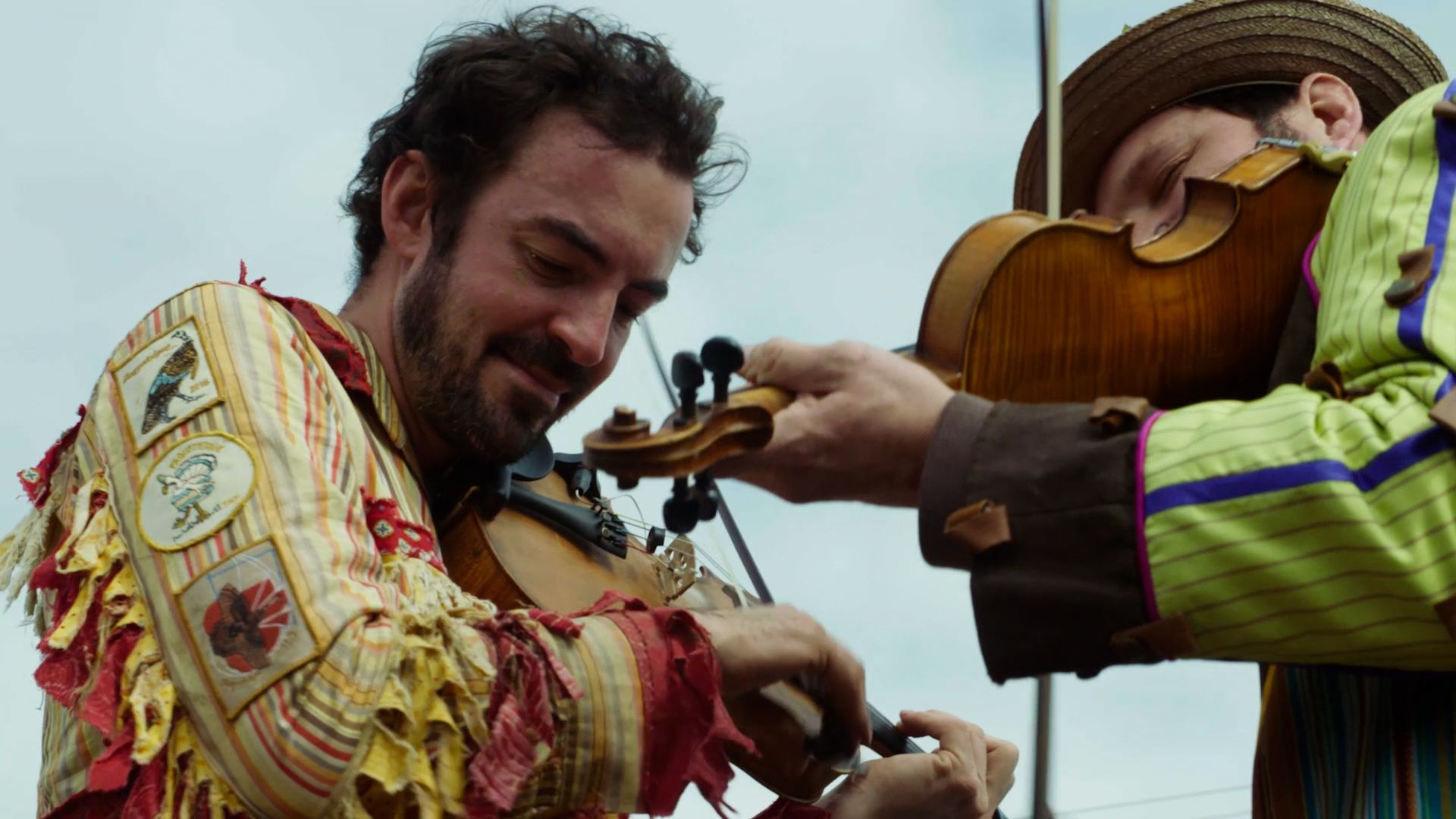 Two men, wearing traditional Cajun Mardi Gras attire, lean toward each other as they each play the fiddle.