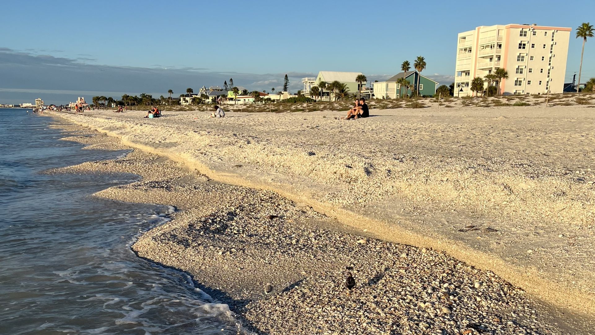 Pebble beach with calm blue water on the left, pastel buildings and palm trees along the shore, and a few people sitting on the sand under a clear sky.