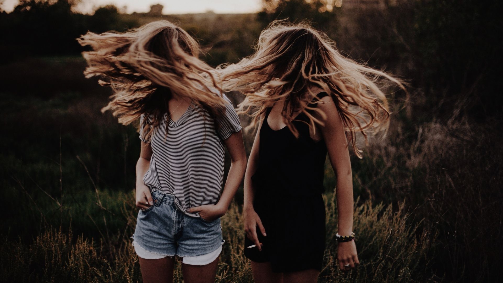 A photo showing two girls with light hair swishing it over their faces in a field. One is wearing a cotton striped V-neck T-shirt and cutoff jean shorts. The other is wearing a black tank and black shorts or it could be a black romper. She is holding a cigarette. 