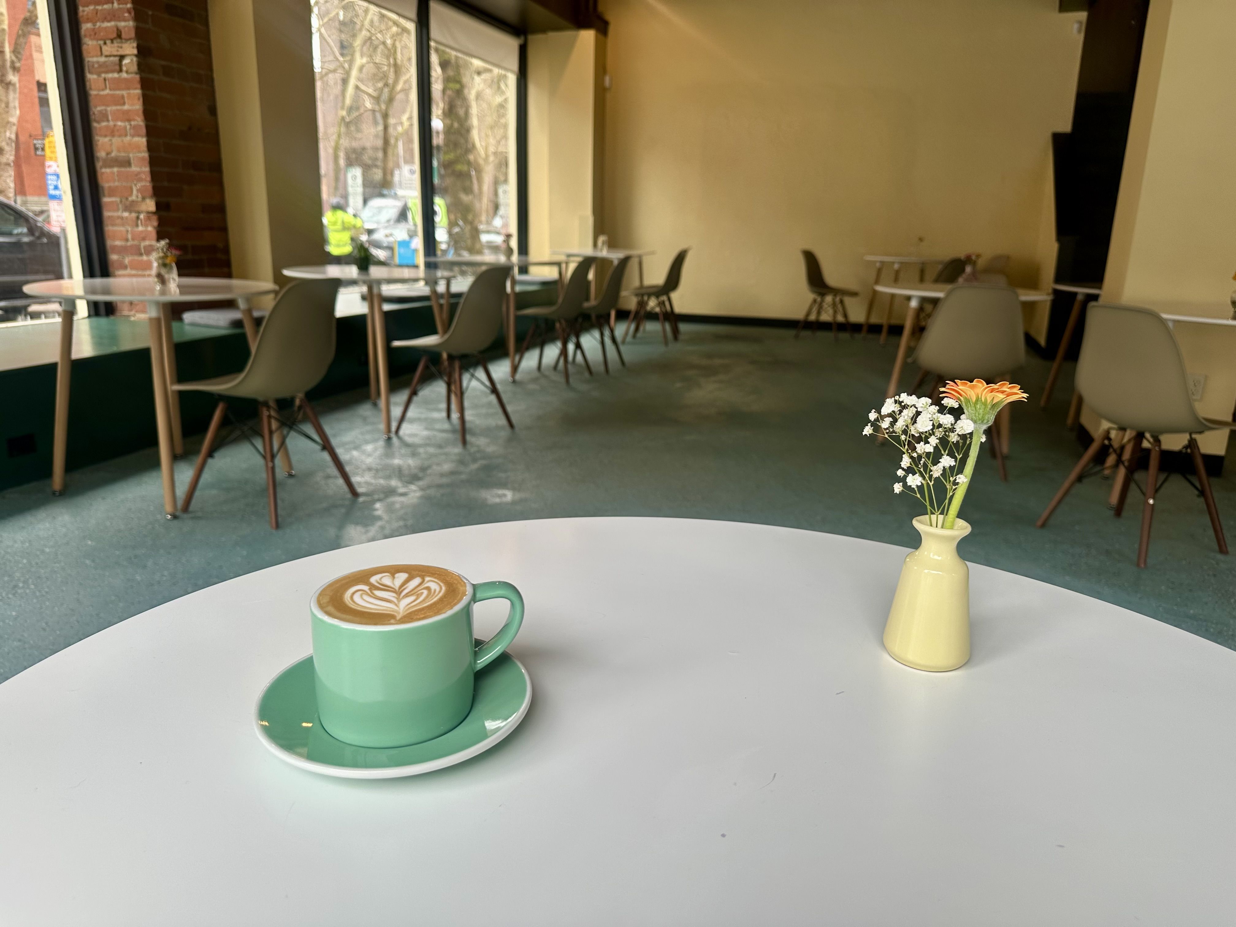 A green coffee cup and saucer with foam art in the form of a flower sits on a white table with a small yellow vase with flours, looking over a industrial chic space with chairs facing a large window.