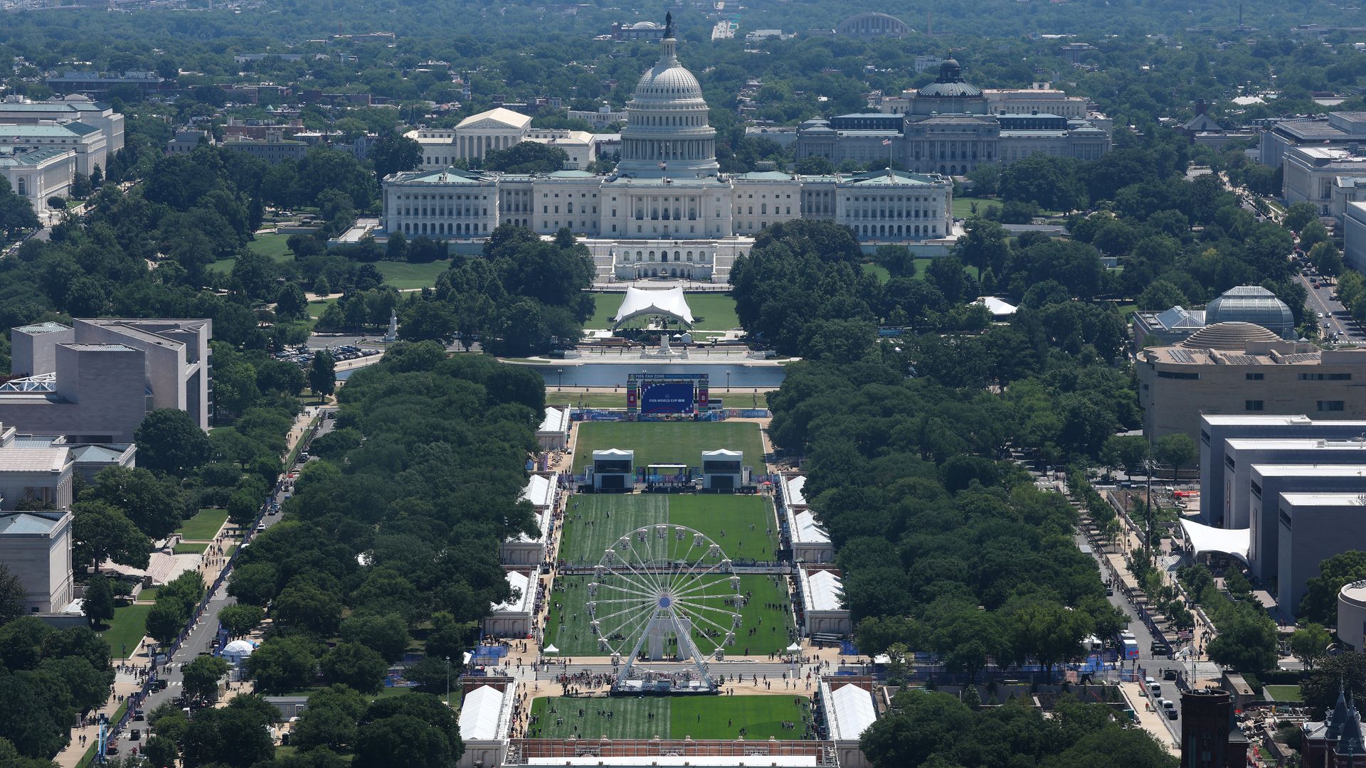 Aerial view of the U.S. Capitol in Washington, D.C., with its dome and wings, surrounded by trees. The National Mall extends forward with green lawns, tents, and a Ferris wheel in the foreground.