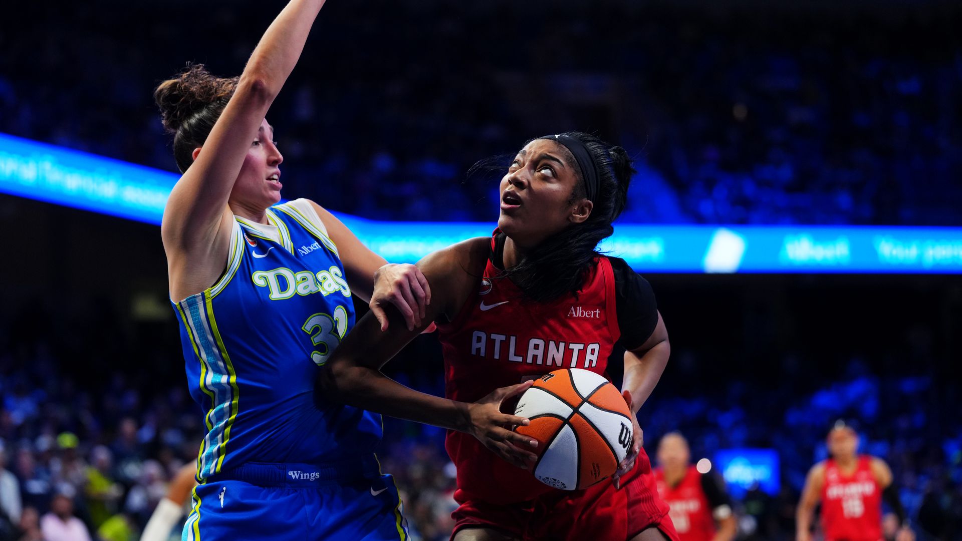 A basketball player wearing a read "Atlanta" jersey pushes their elbow against an opposing player wearing a blue "Dallas" jersey and matching shorts.
