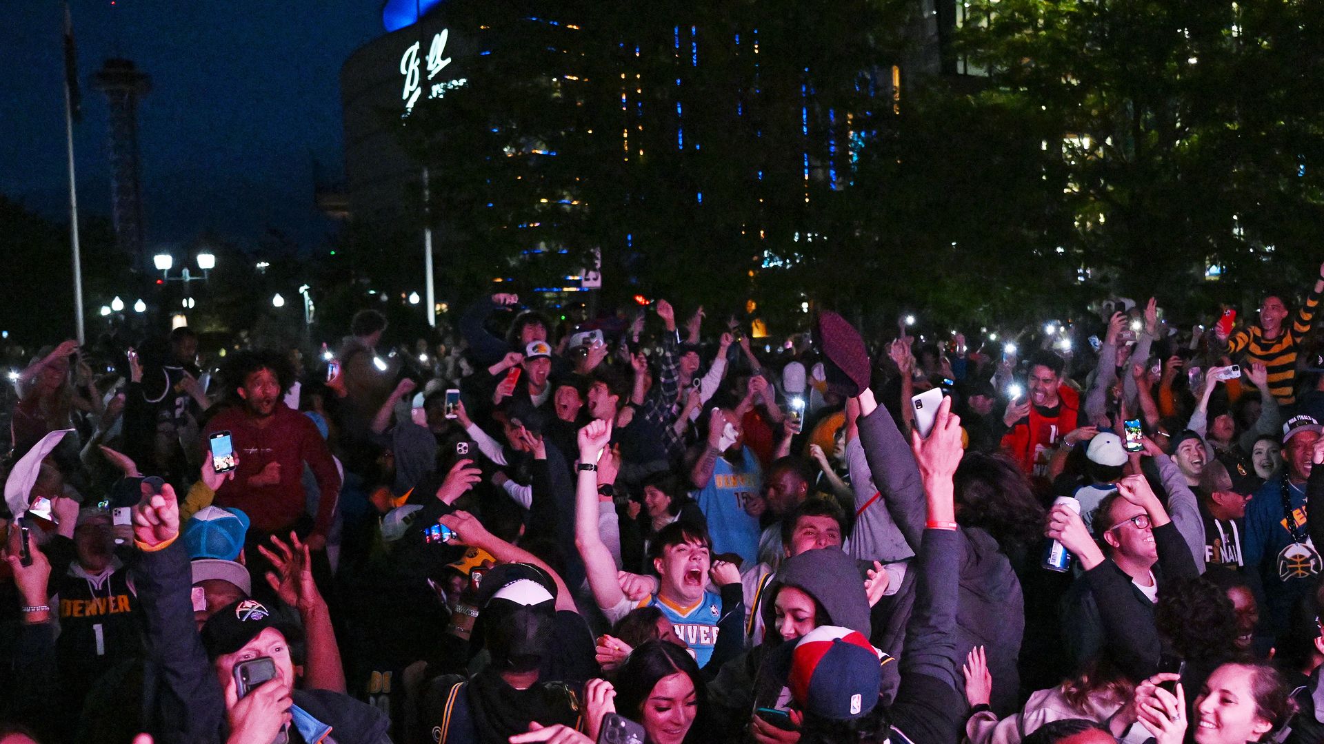 Nighttime crowd of excited fans cheering, holding up smartphones with lights. Many wear sports jerseys; city lights and a tall building glow in the background.