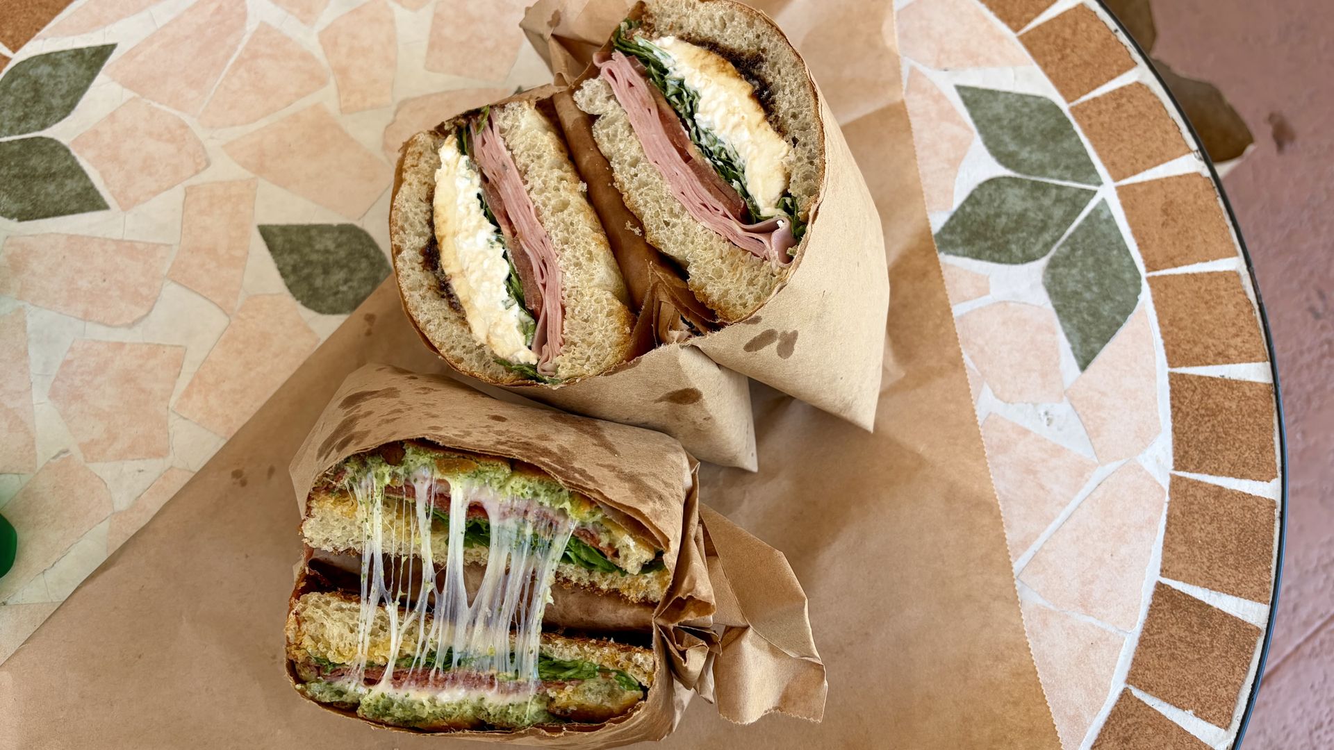 Two deli sandwich halves on a mosaic round table with brown paper wrappers. 