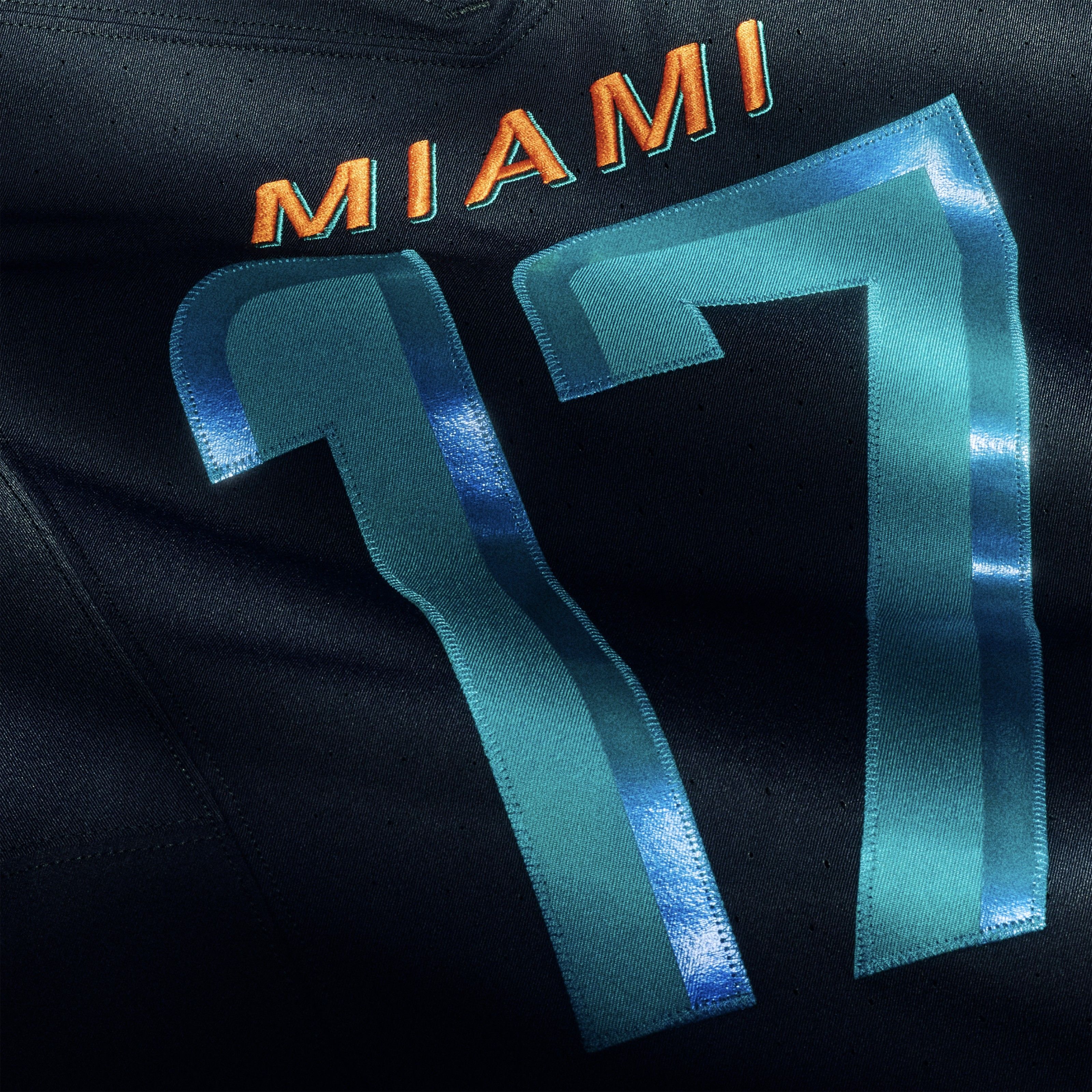 Black fabric with bright orange text "MIAMI" above large blue and teal number "17" stitched onto it, showing a sports jersey detail.