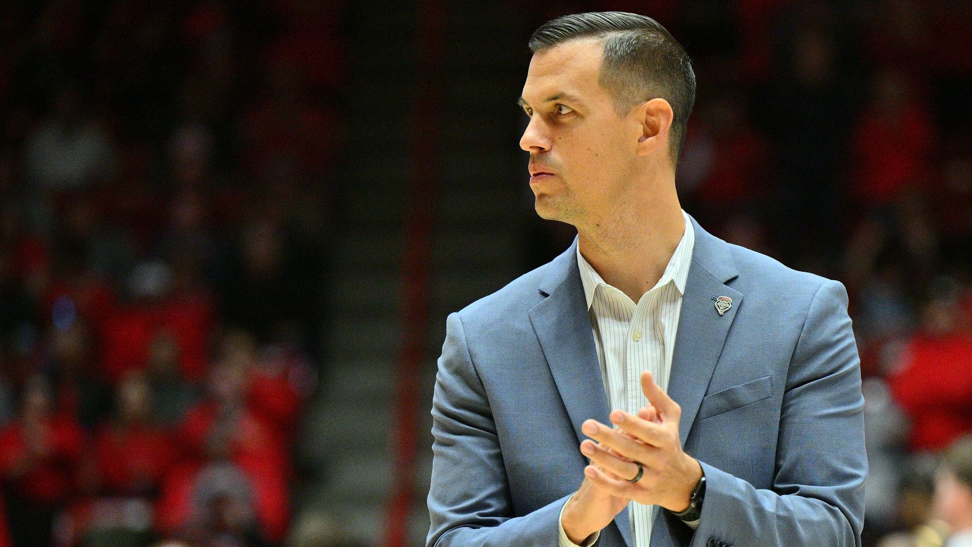 Fernando Lovo in a blue suit jacket and white shirt clapping hands, looking to the side, indoors with blurred crowd in red and dark background.