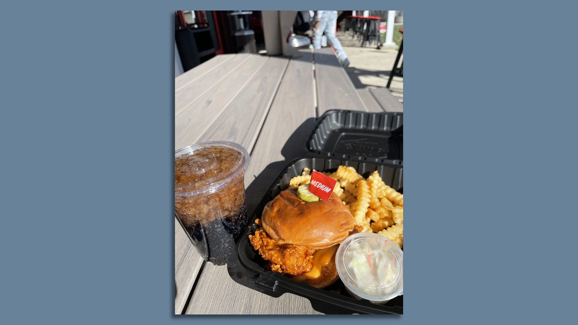 Takeout meal on a picnic table with a fried chicken sandwich labeled medium spice, crinkle-cut fries, coleslaw, and a plastic cup of soda with ice outdoors on a sunny day.