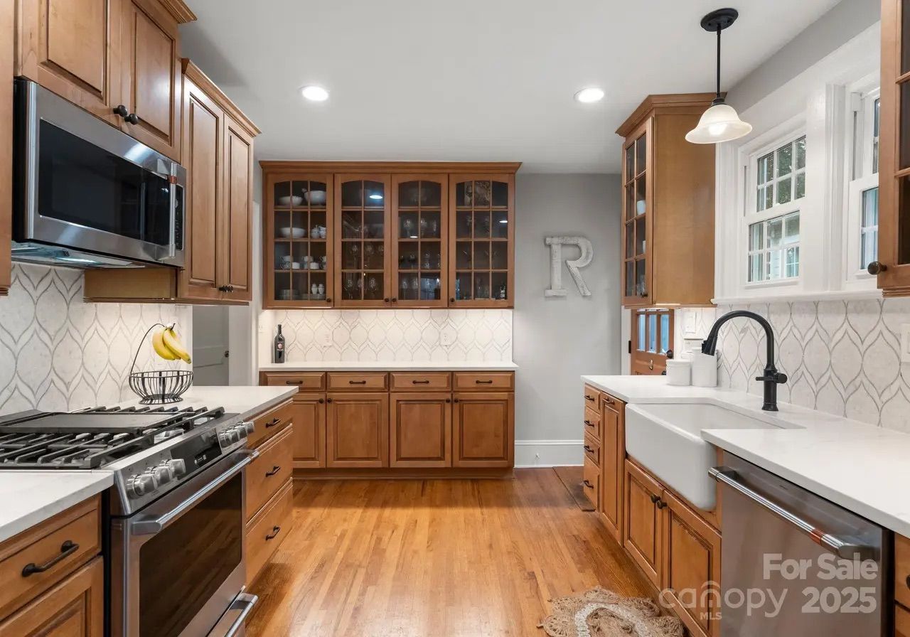 Bright kitchen with wooden cabinets, white countertops, farmhouse sink, stainless steel stove and microwave, patterned backsplash, hardwood floor, and wall letter "R".