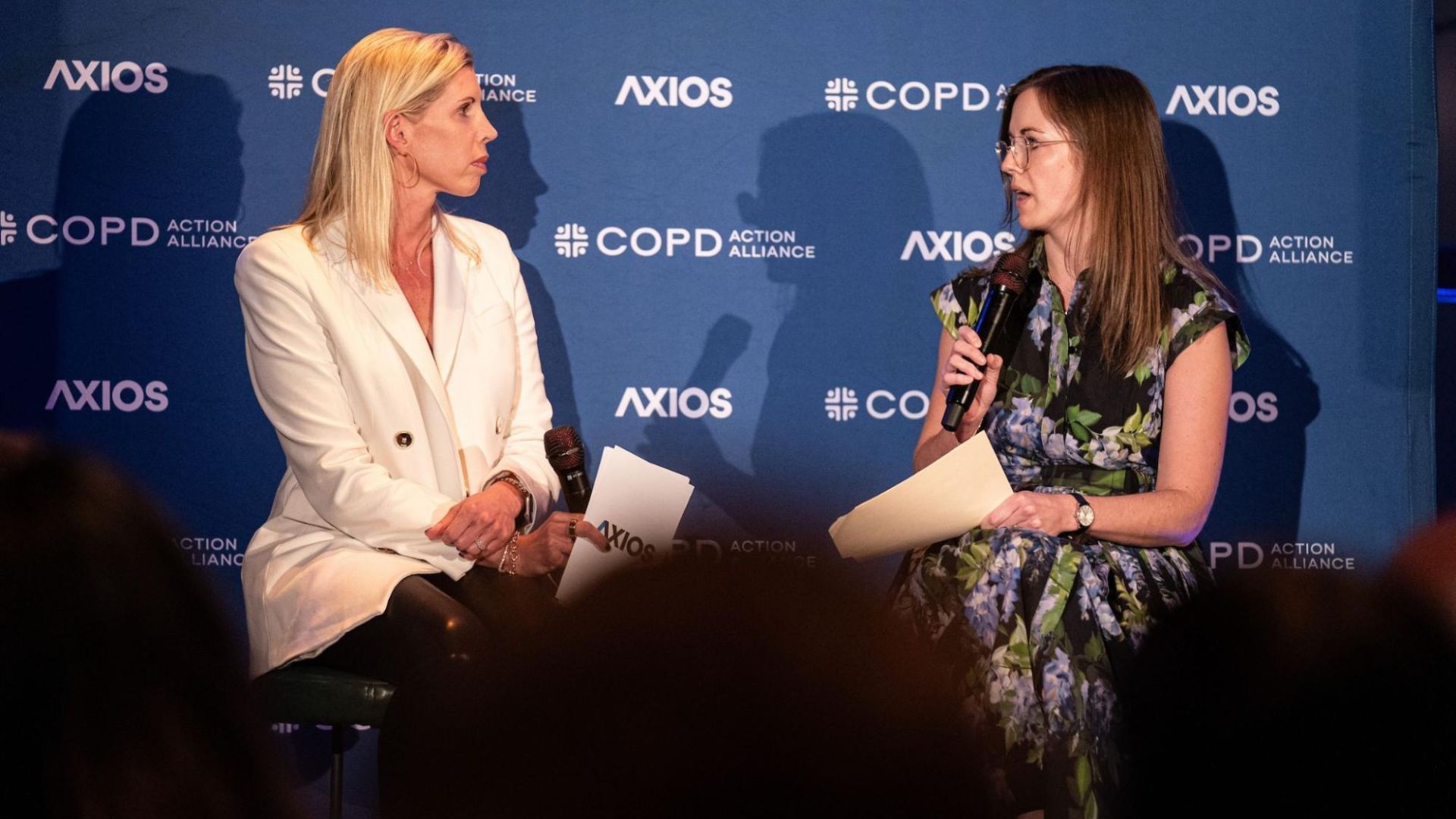 Two women with microphones seated on stage during a discussion, one in a white blazer and the other in a floral dress, against a blue backdrop with Axios and COPD Action Alliance logos.