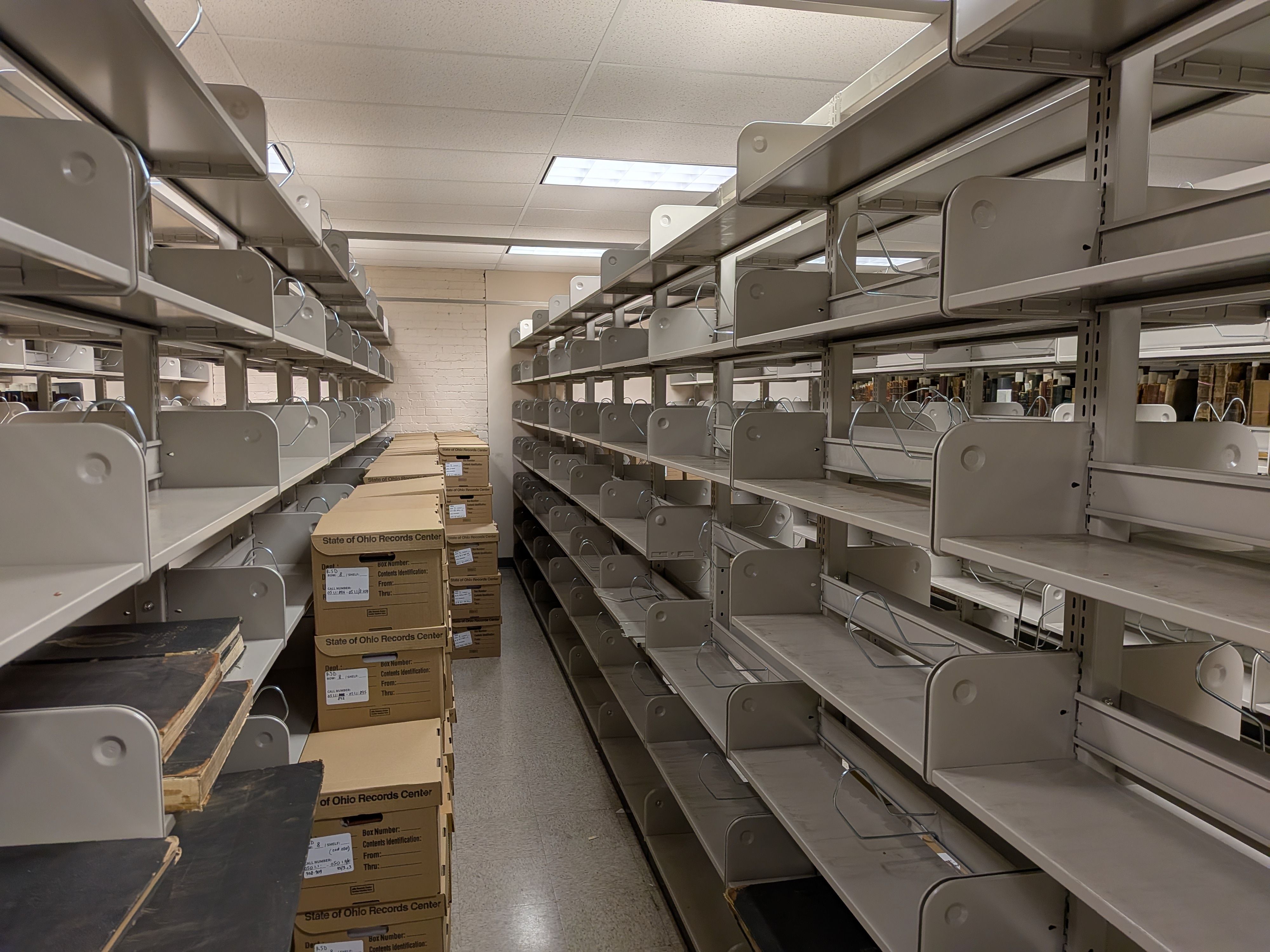 Storage room with long rows of gray metal shelves on both sides, forming an aisle. Empty shelves and stacked brown boxes labeled "State of Ohio Records Center" line the floor under fluorescent lights.