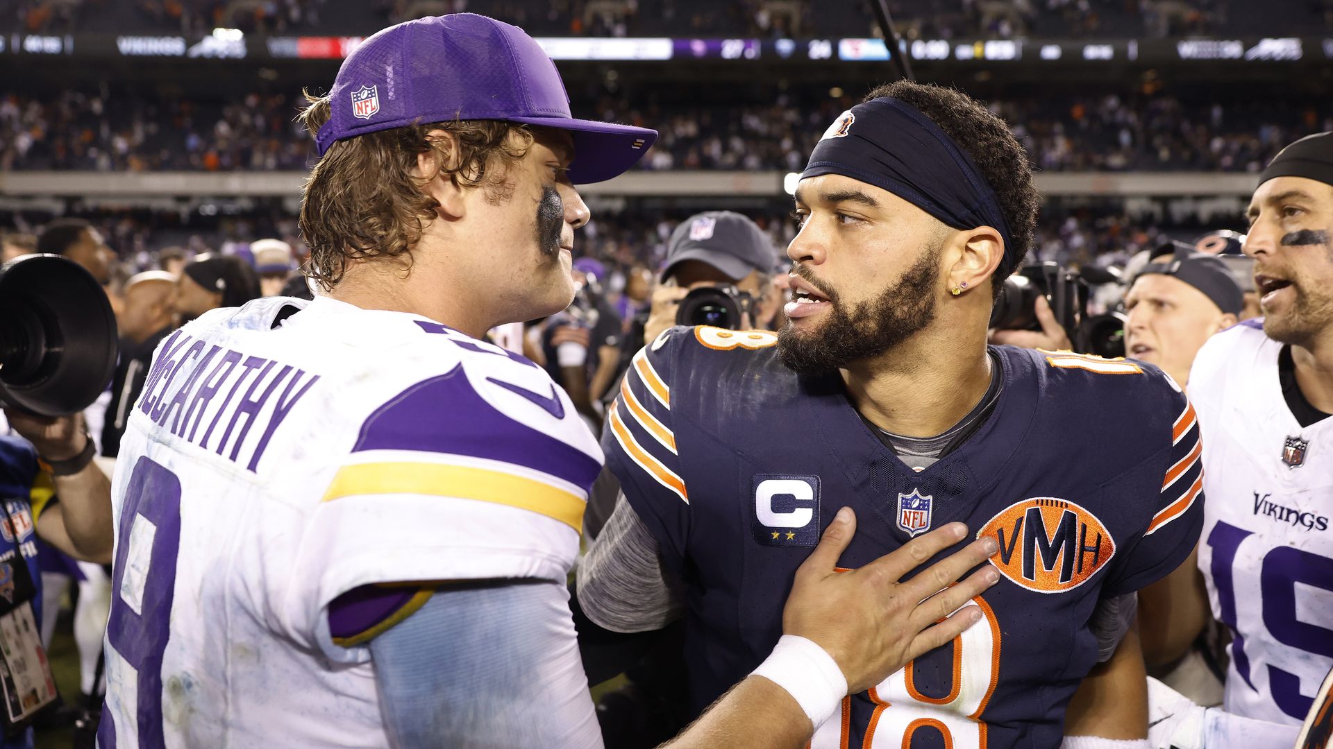 JJ McCarthy, wearing a white jersey and purple hat, taps the chest of Caleb Williams, who is wearing a dark blue jersey. Adam Thielen is the background. 