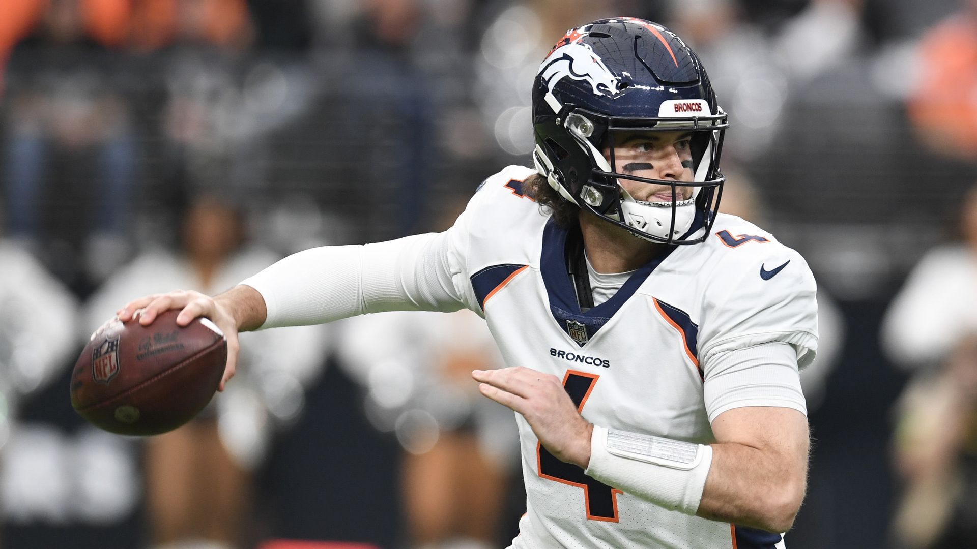 A man wearing a navy blue helmet and white jersey with orange lining prepares to throw a football with his right hand during an American football game in Las Vegas. 