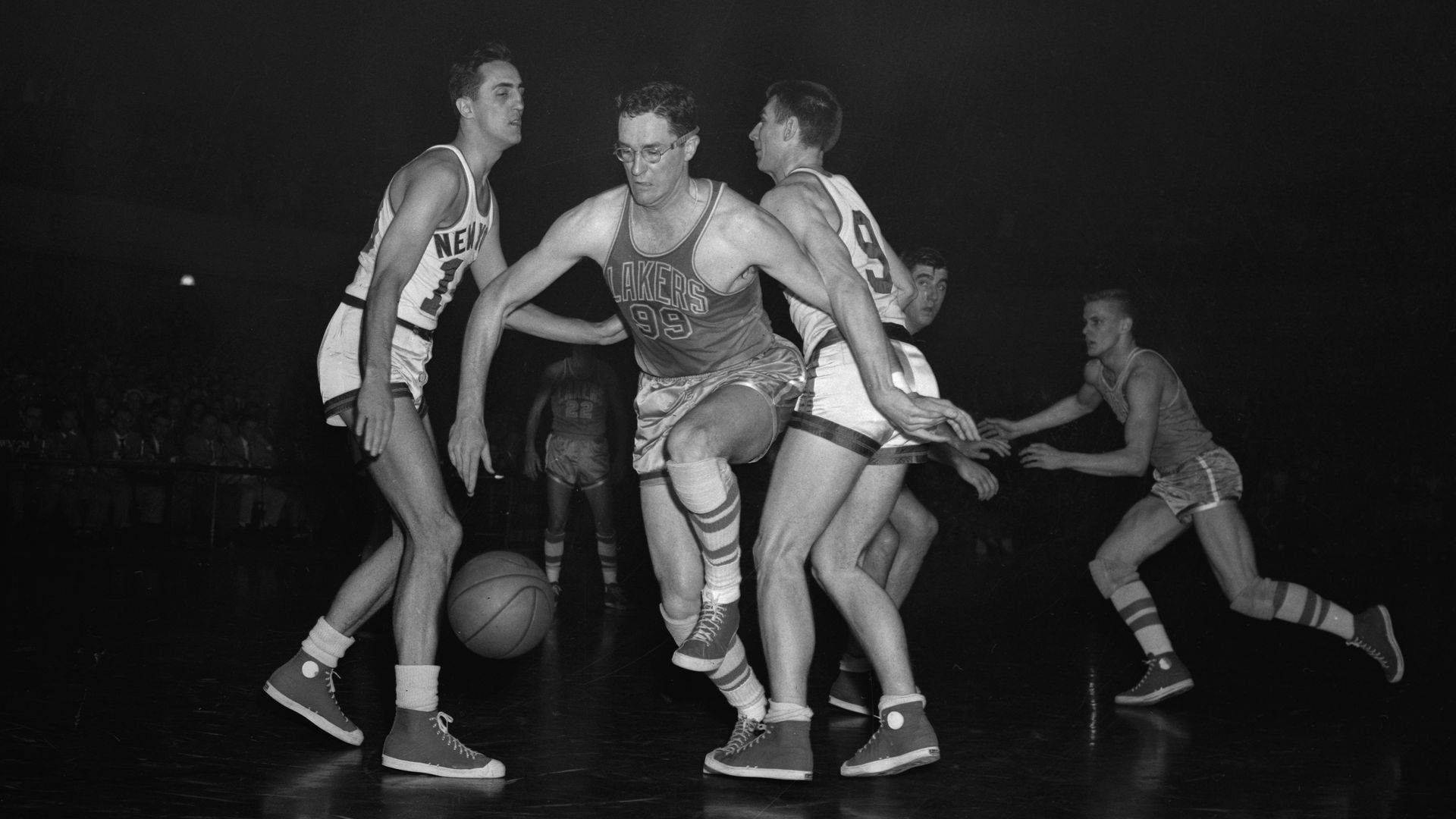 Black and white photo of a basketball game with players in vintage uniforms, one with glasses dribbling the ball near the basket, scoreboard showing 20-26 in the background.
