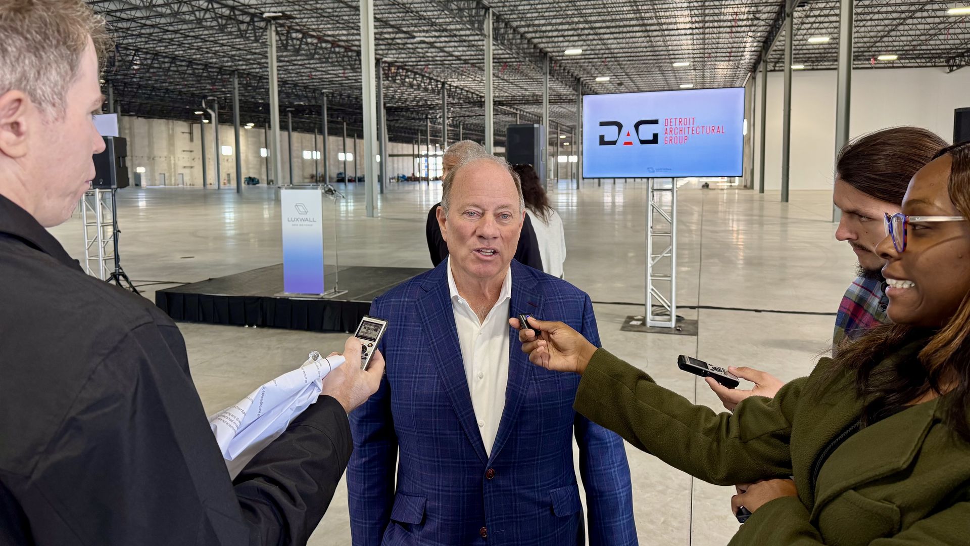 Duggan speaks in center frame with three reporters crowded around recording his answers. He's standing in an empty manufacturing facility.