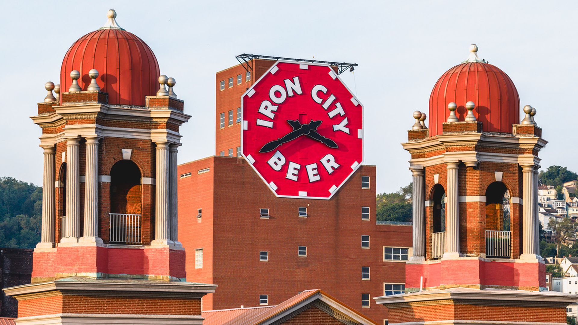Two red domed towers with white columns frame a large red octagonal sign on a brick building that reads "IRON CITY BEER" with clock hands in black.