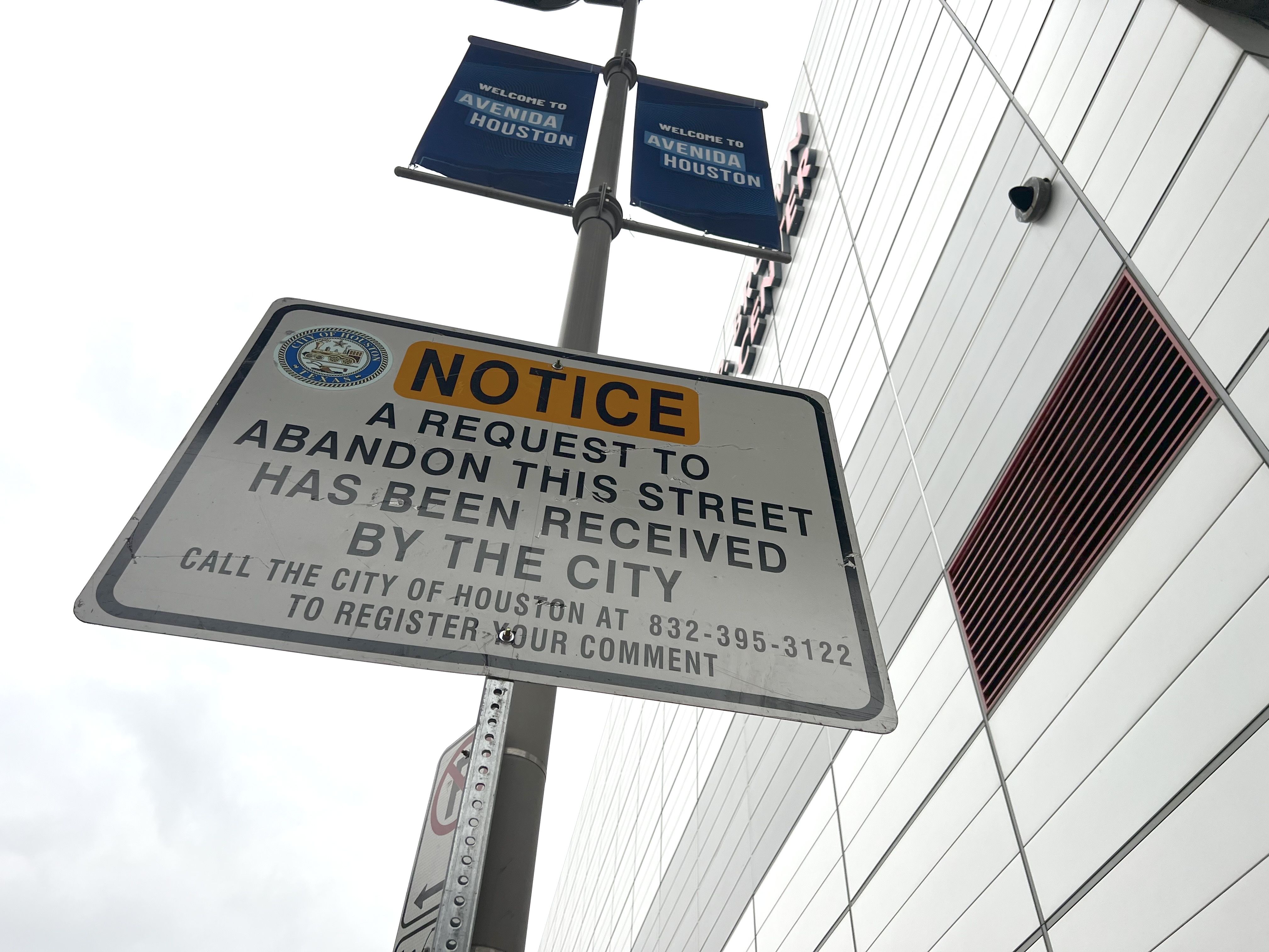 The George R. Brown Convention Center looms over a sign that reads "NOTICE A request to abandon this street has been received by the city. Call the city of Houston at 832-395-3122 to register your comment"