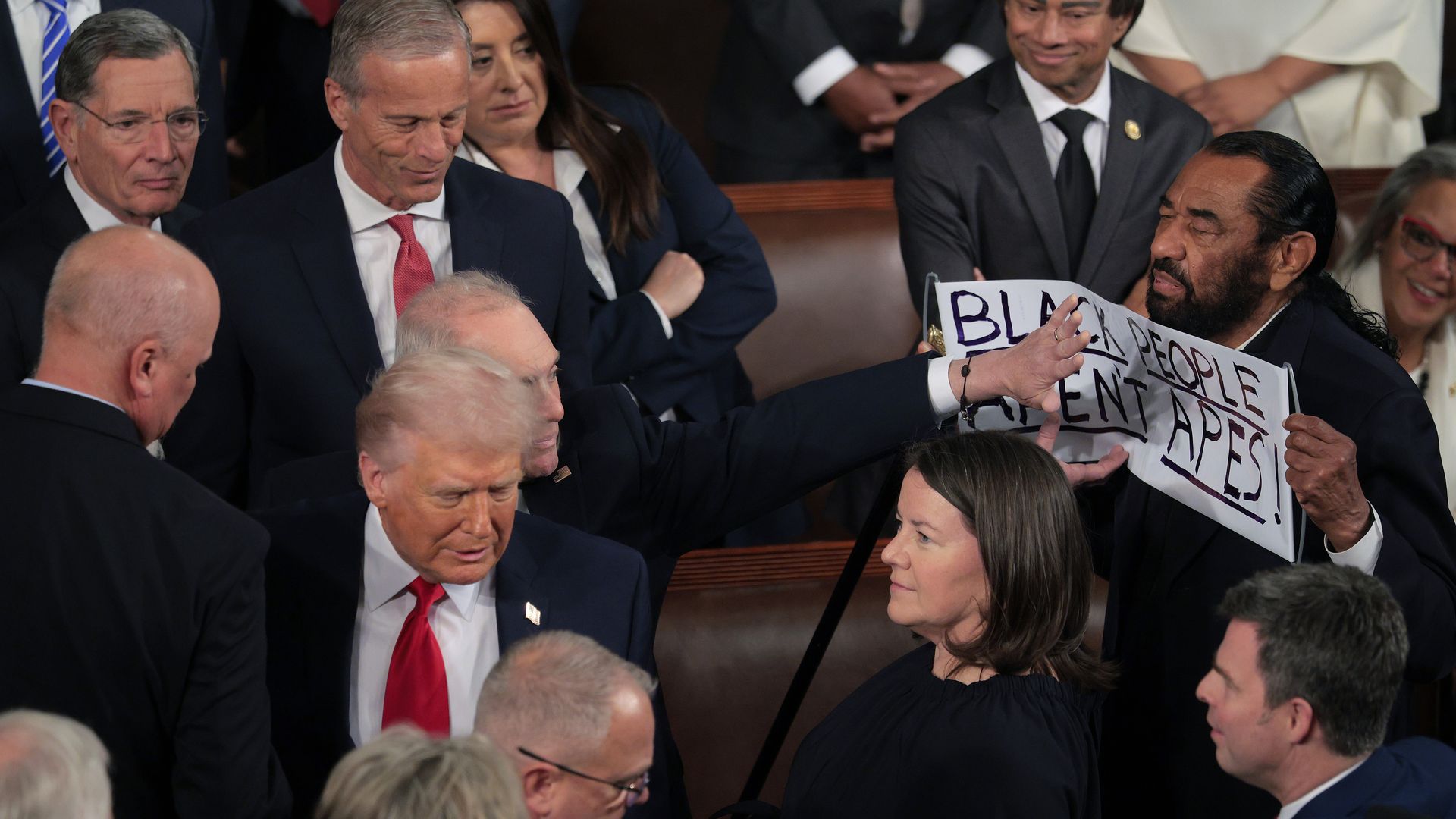 Man holding a sign reading "BLACK PEOPLE AREN'T APES!" is stopped by a man with outstretched arm in a crowded room of formally dressed people, including one man in a red tie.