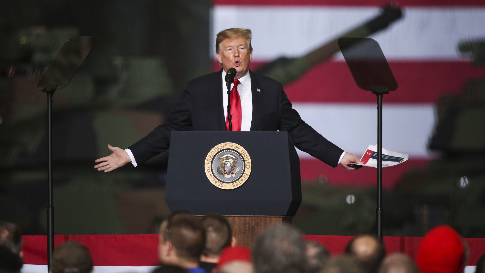 Trump stands at a podium while holding papers in one hand, with the American flag behind him. A crowd watches.