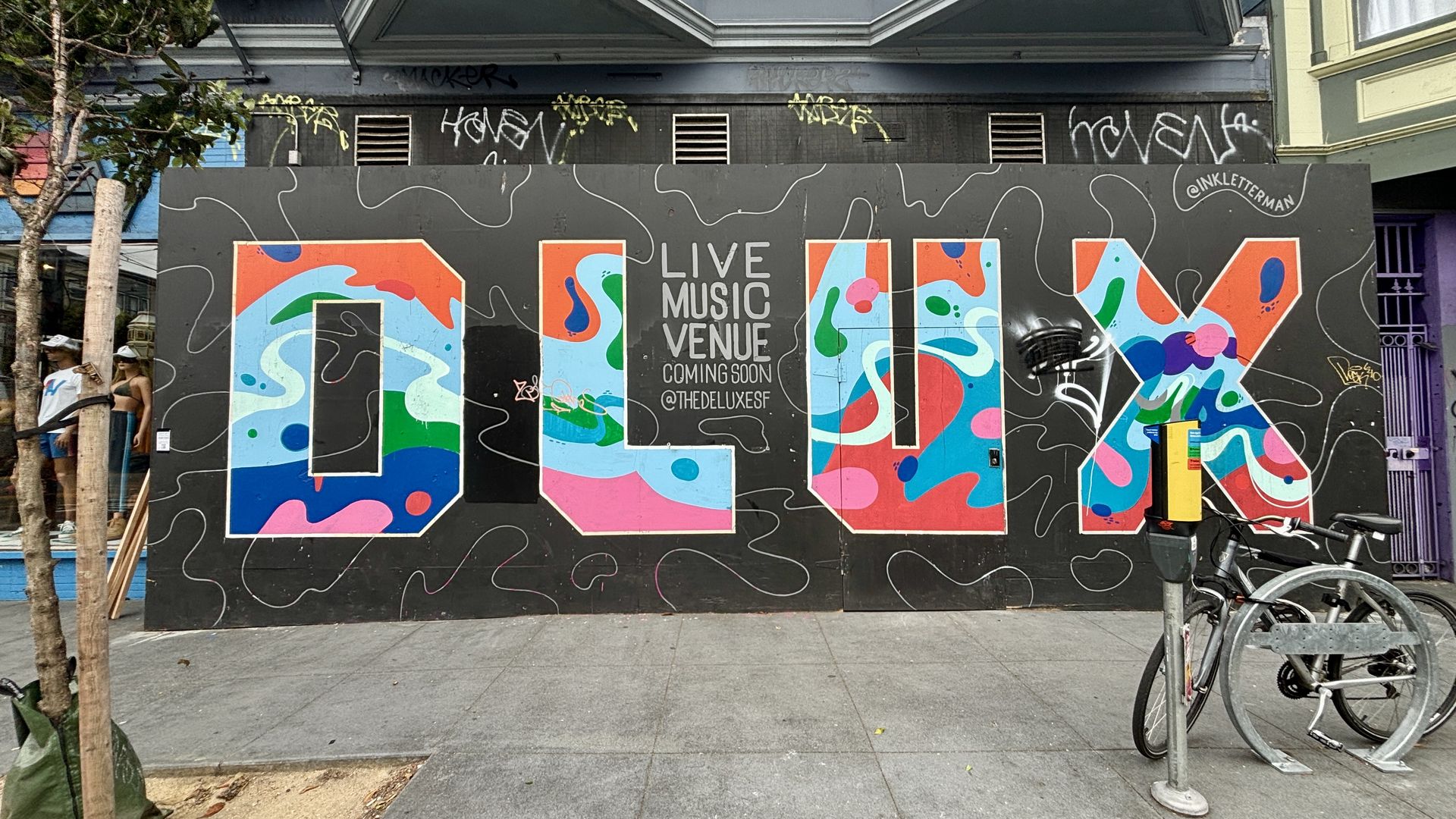 Gray wall mural with large colorful letters spelling "LOVE" filled with abstract shapes. Center reads "LIVE MUSIC VENUE COMING SOON @THEDELUXESF". Left: two people by a tree; bikes on the right.