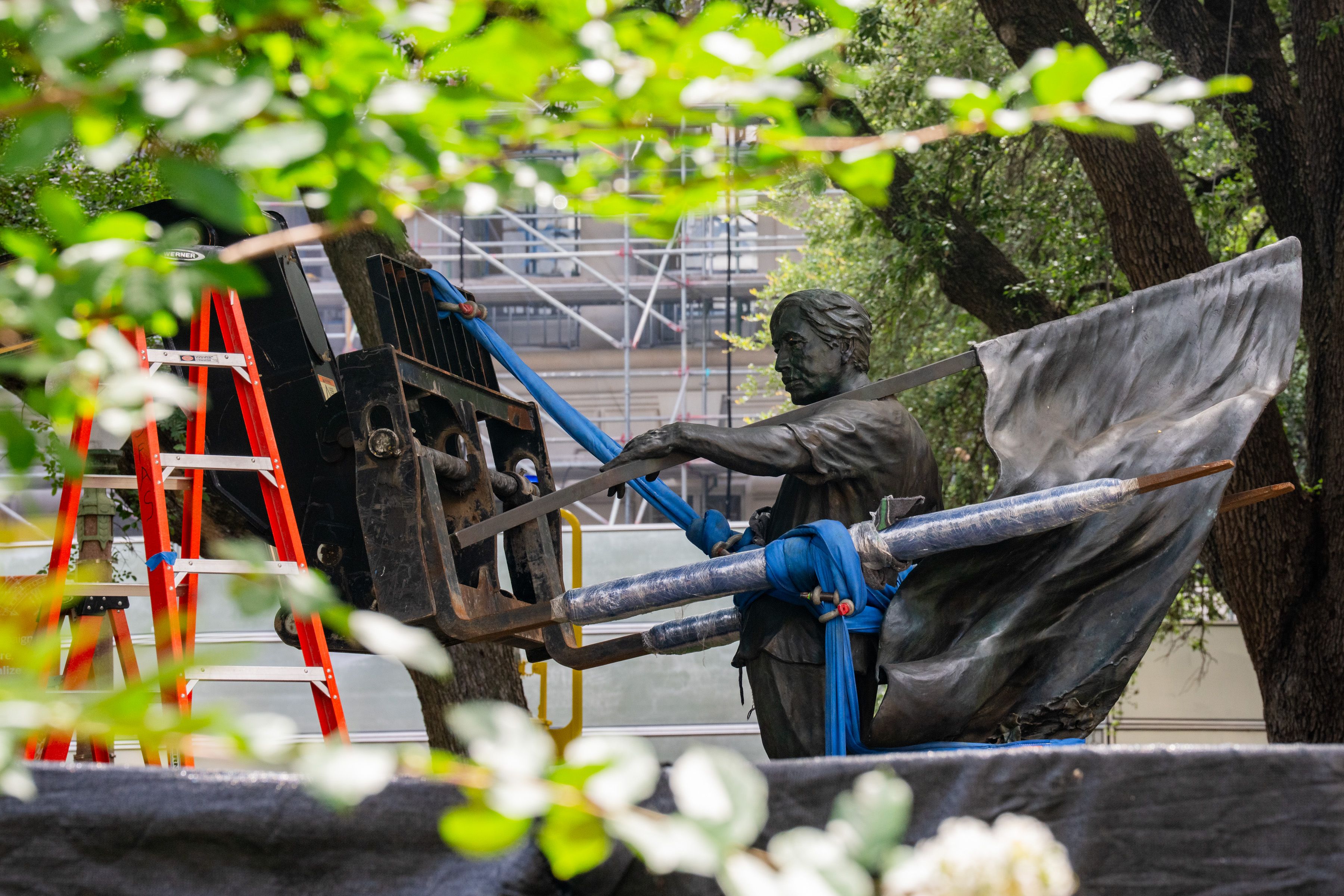 Bronze statue of a man reaching forward, gripping a blue strap attached to a large black construction vehicle. Red ladders, scaffolding in the background, with green tree foliage in front.