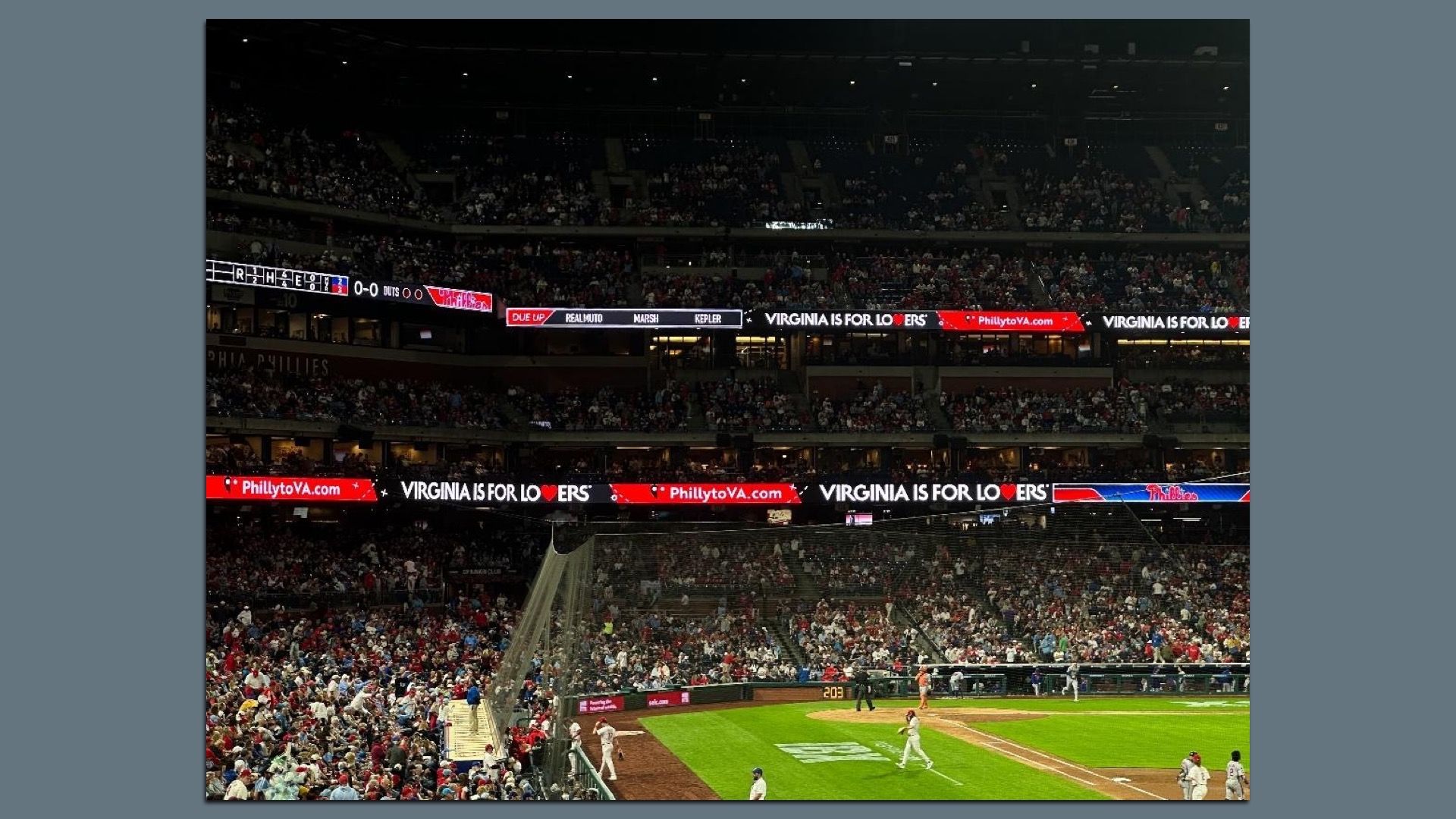 A crowded baseball stadium at night with fans in the stands. Outfield LED boards read "Virginia is for Lovers" and "PhillytoVA.com"; players on the green field near home plate.