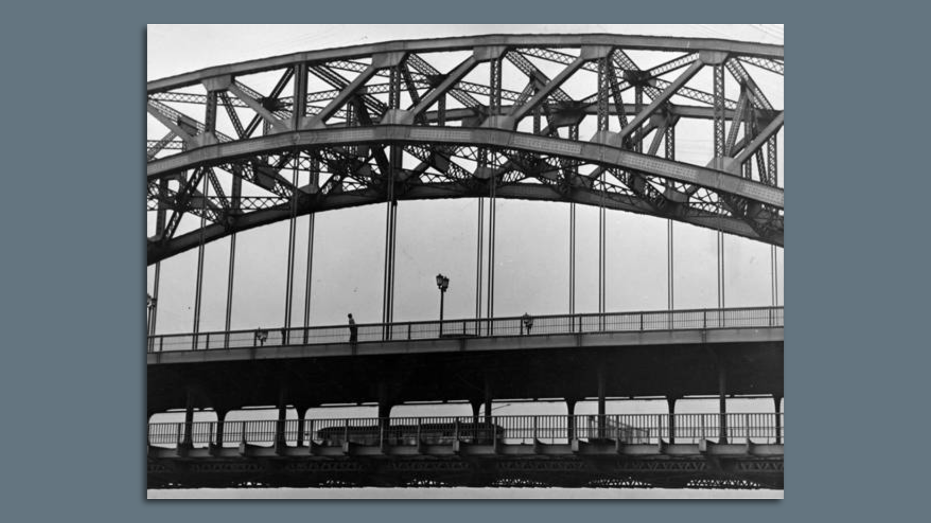 A black and white image of a streetcar traveling under a suspension bridge.