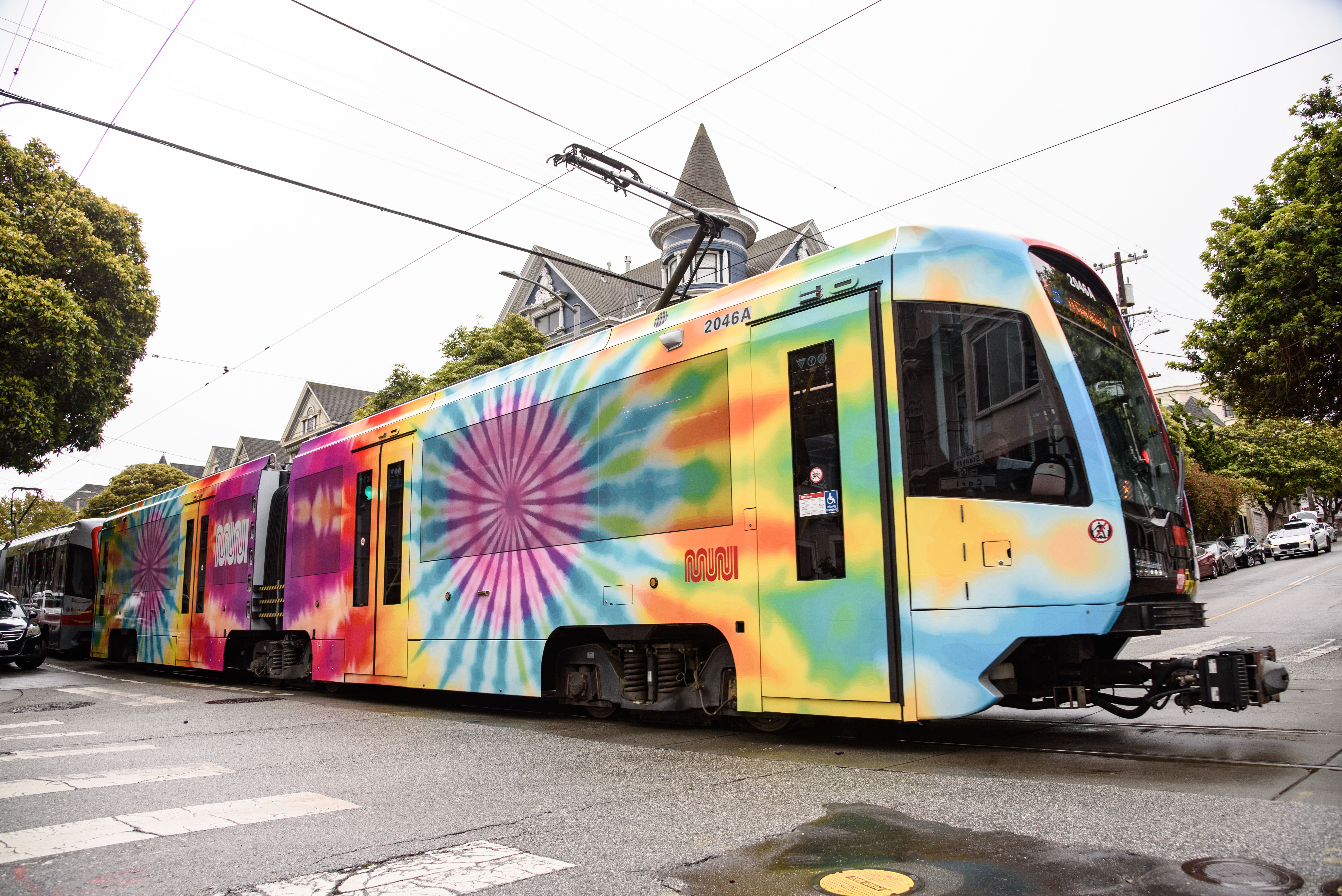 Colorful streetcar with tie-dye pattern in shades of pink, green, blue, yellow, and orange, traveling on city street with Victorian-style houses and green trees.