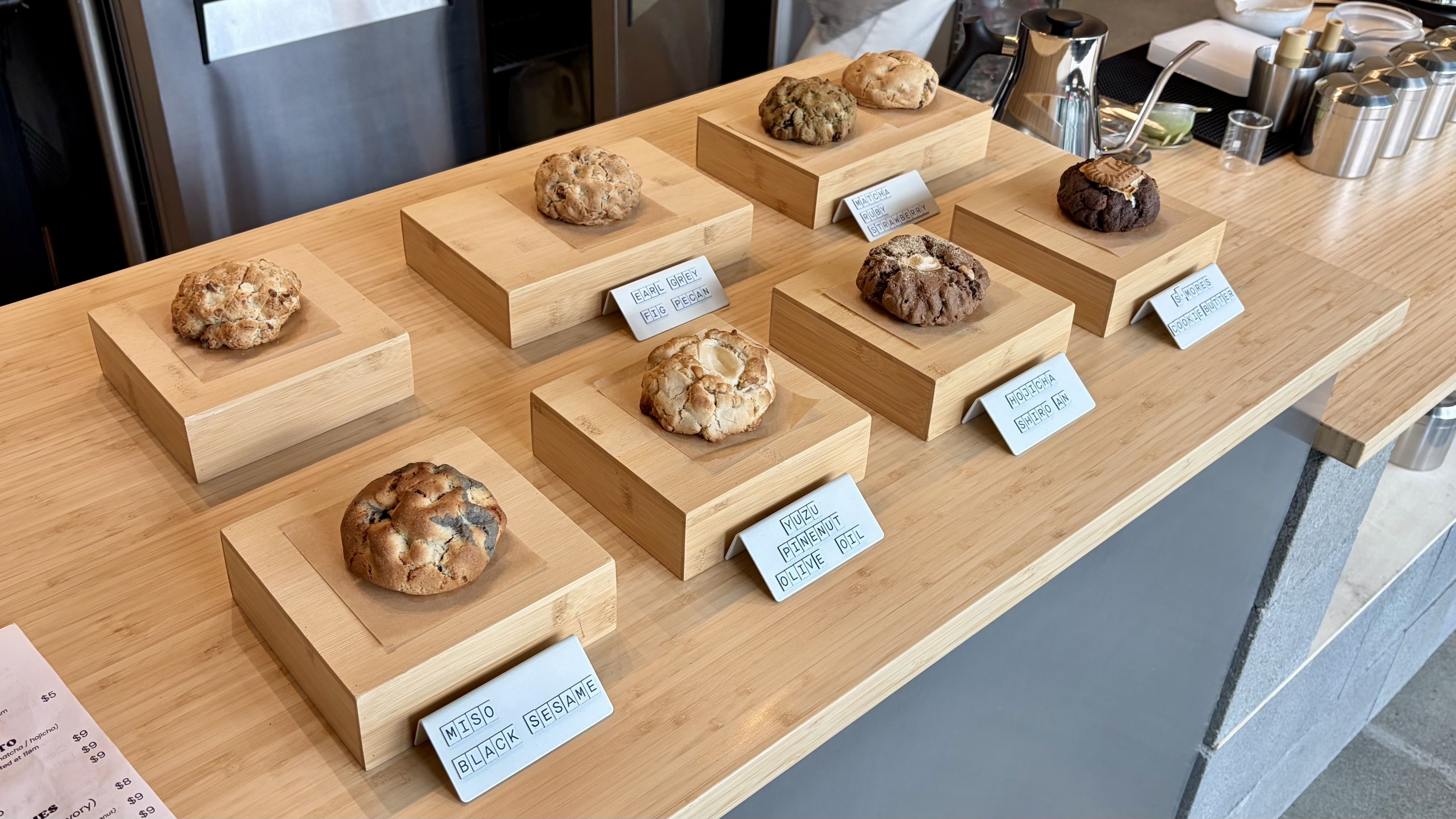 Cookies on light wooden pedestals arranged along a cafe counter; cookies range from light to dark brown with varied textures. Small white cards list flavors like Earl Grey Fig Pecan and Matcha Ruby Strawberry.