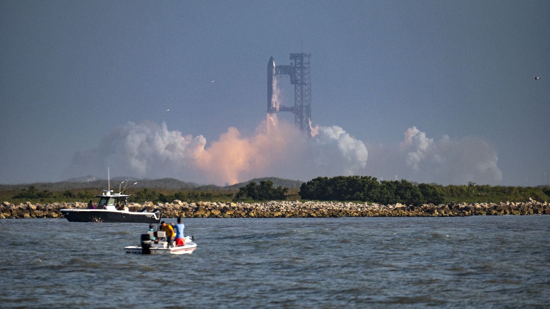 A rocket launches amid lots of vaporous clouds in the distance as two boats watch from the water. 