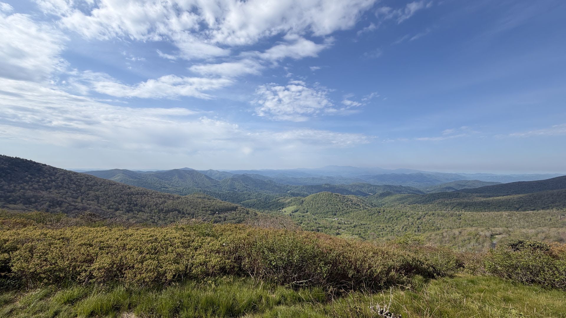 Panoramic mountain landscape under a blue sky with white clouds. In the foreground, green grass and brown shrubs lead to rolling ridges and distant hills fading to the horizon.