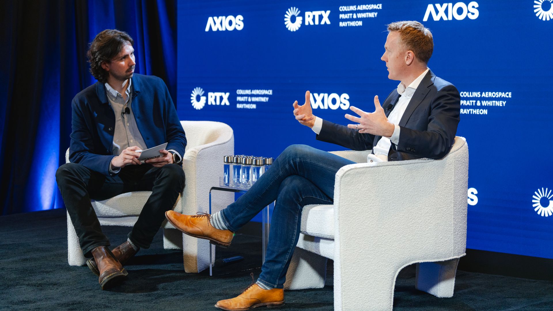 Two men in business casual sit onstage at an event. The chairs are white. The backdrop is dark blue.