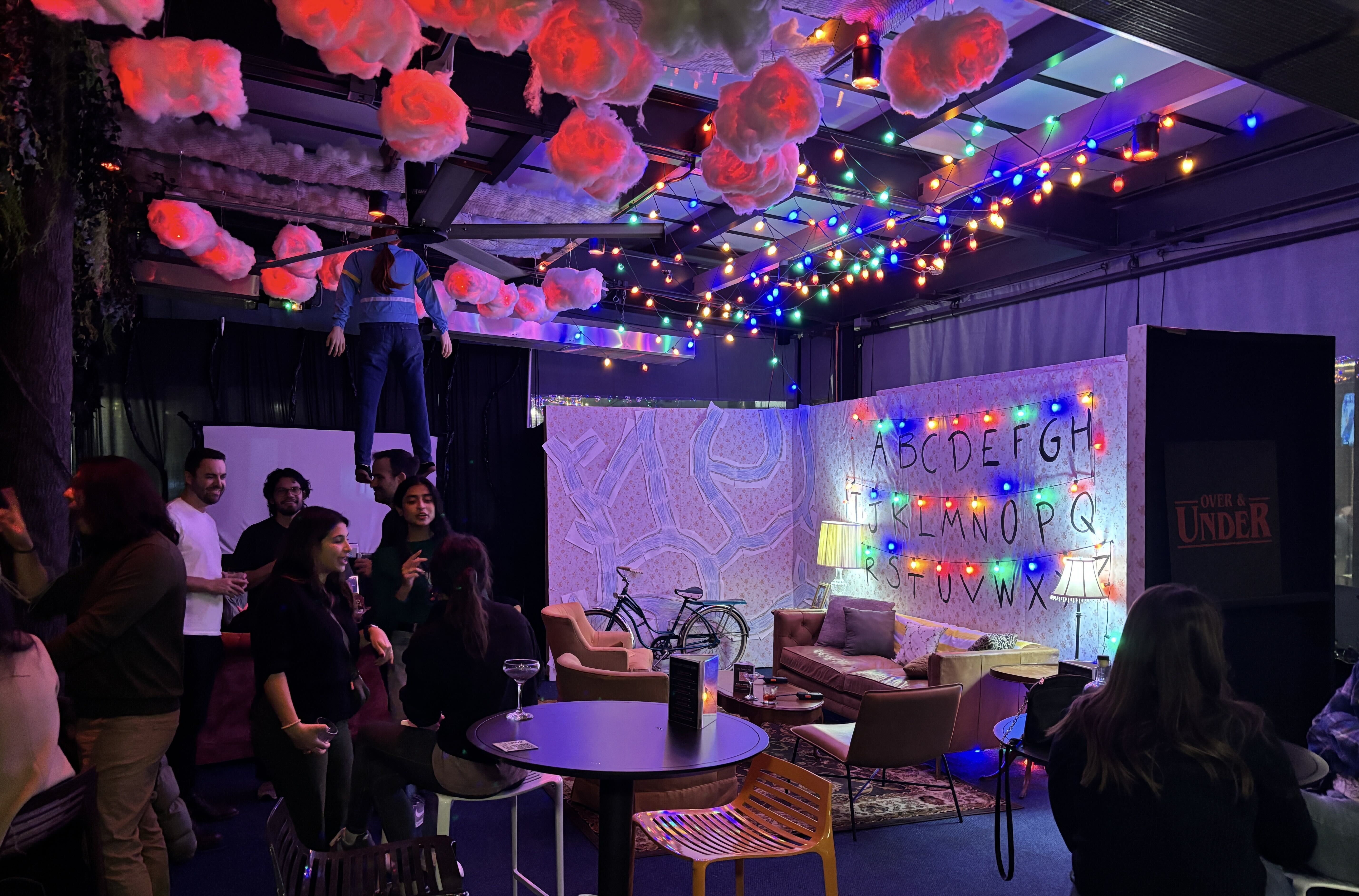 Dimly lit room decorated with glowing red clouds on ceiling, colorful string lights, a wall with alphabet letters, cozy seating areas, and people socializing in a casual indoor setting.