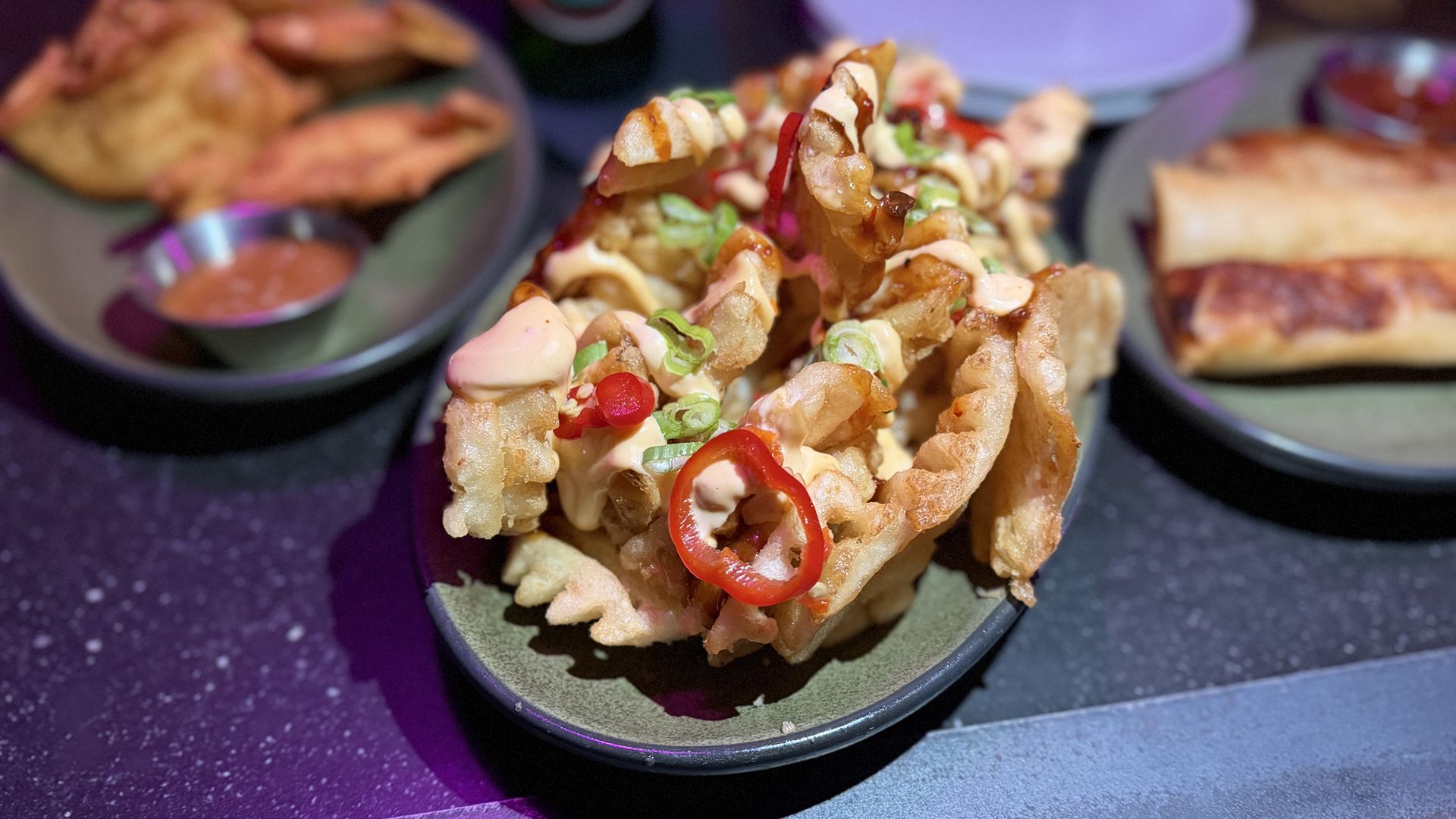 Plate of waffle fries topped with sliced red chili, green onions, and creamy sauce, surrounded by plates with fried appetizers and dipping sauce on a dark table.