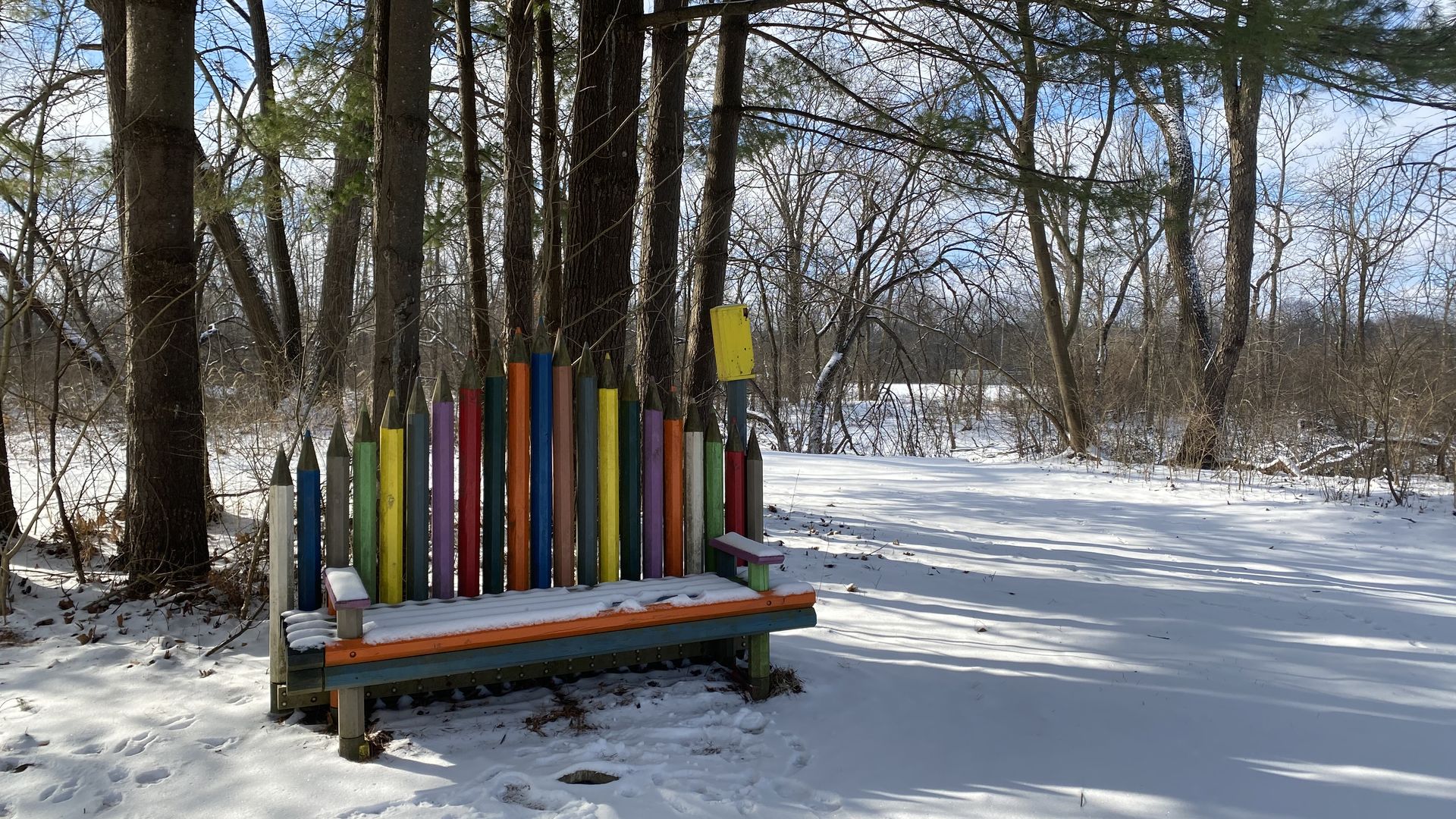A park bench made out of colored pencil shaped wood in a snowy park near trees