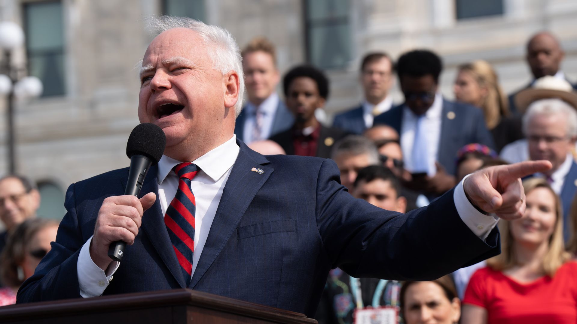 A man in a blue suit with a white dress shirt and blue-and-red-striped tie holds a microphone and points while giving an emphatic speech