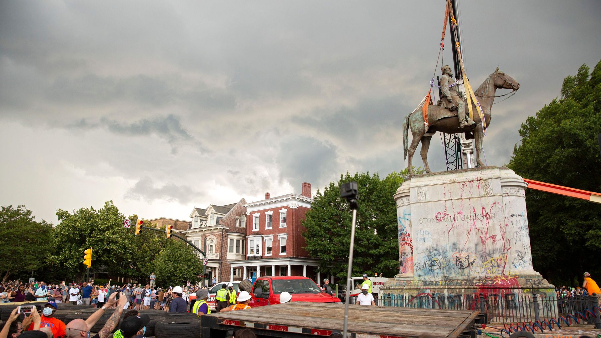 A picture of a crowd looking at a Confederate general and his horse being removed. The confederate is on a graffitied pedestal.