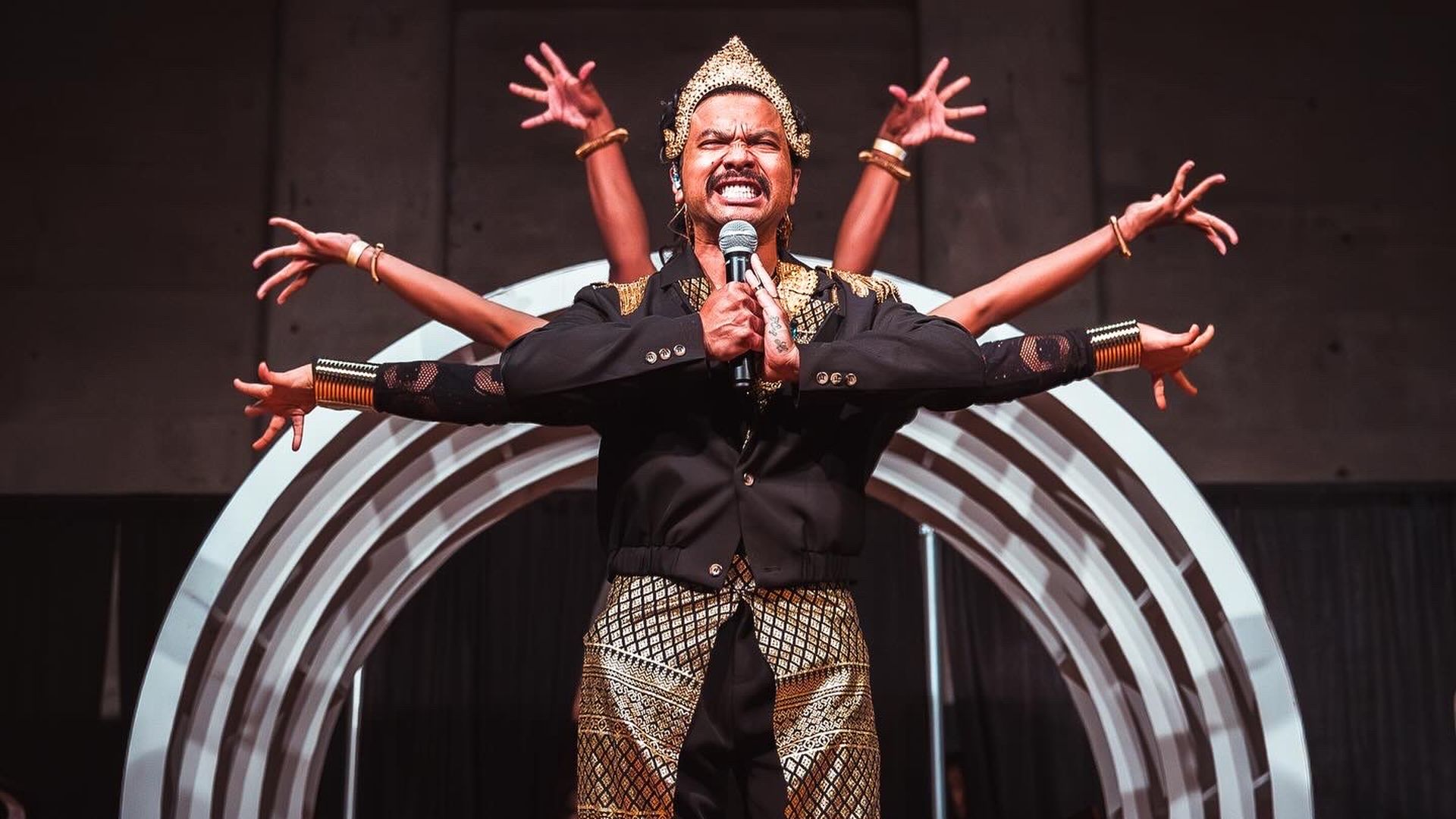 A male performer in a black jacket and gold patterned pants sings into a mic, wearing a gold headdress. Multiple arms extend from behind him, with a metallic circular arch on stage.