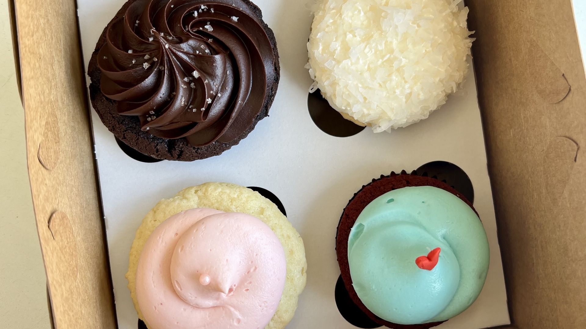 Four assorted cupcakes in a brown box: chocolate with dark frosting and salt flakes, coconut with white flakes, vanilla with pink frosting, and red velvet with light blue frosting and a small red heart.
