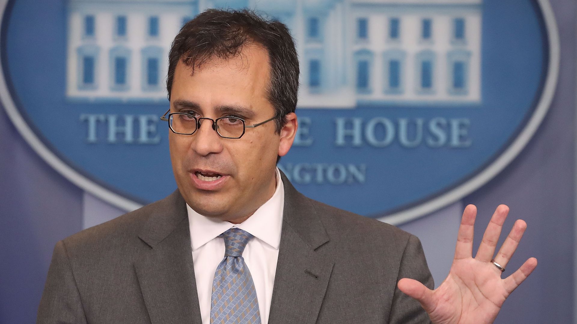 Man with glasses holds up left hand in front of a white house sign