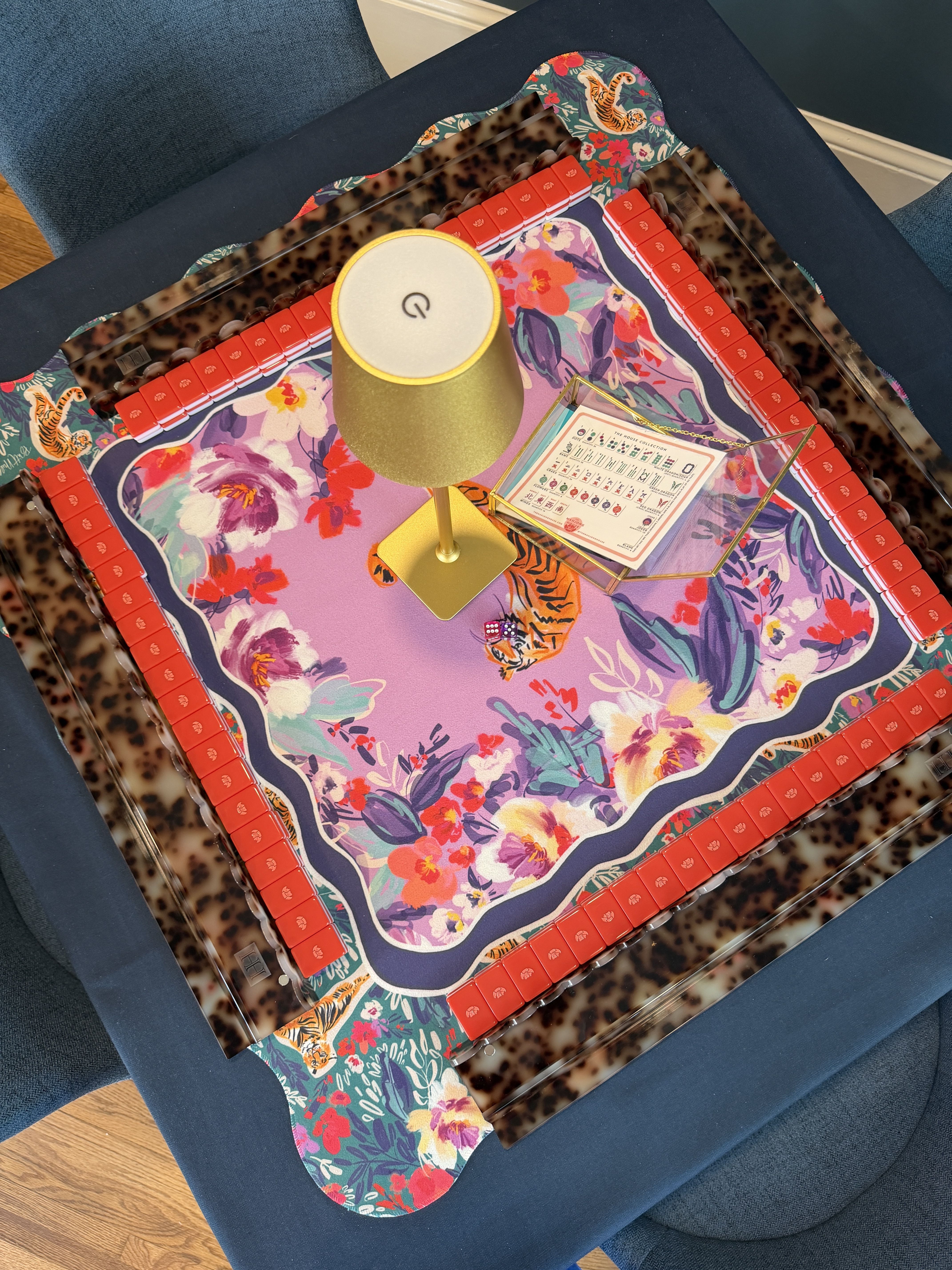 Colorful decorative game board on a dark blue sofa with a pink-purple floral center, red tile border, leopard print background, and a gold cone lamp with a white round top resting on it.