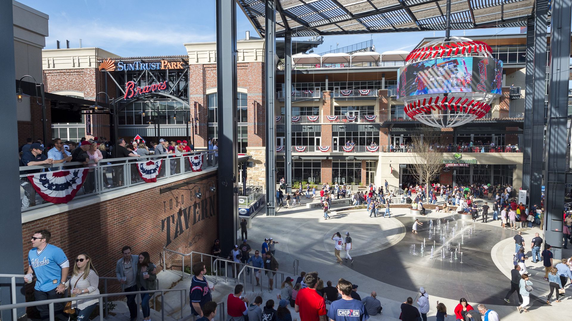 Crowd of people at SunTrust Park, home of the Atlanta Braves, with red, white, and blue bunting, a large baseball-shaped digital screen, and a water fountain in the plaza.