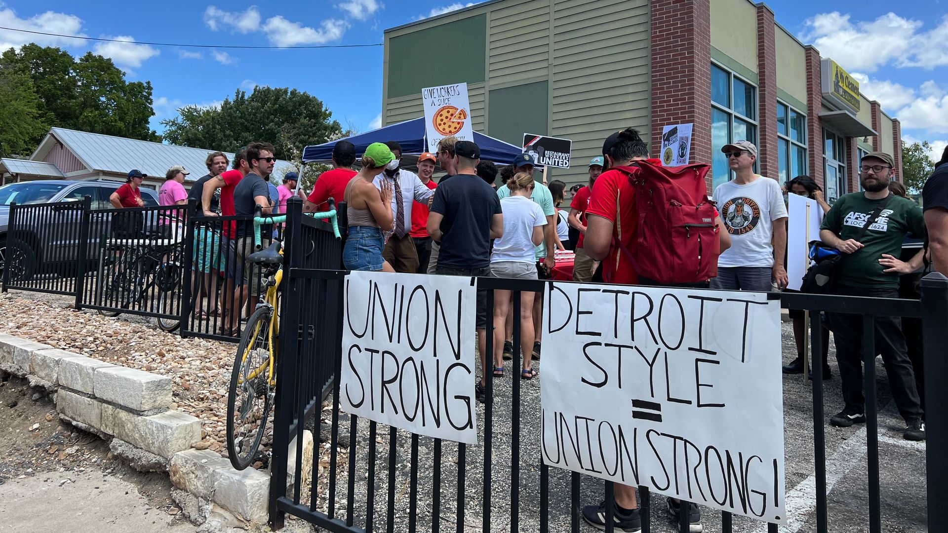 People gather outside with two signs that read "union strong" and "Detroit style=union strong" on a black fence.