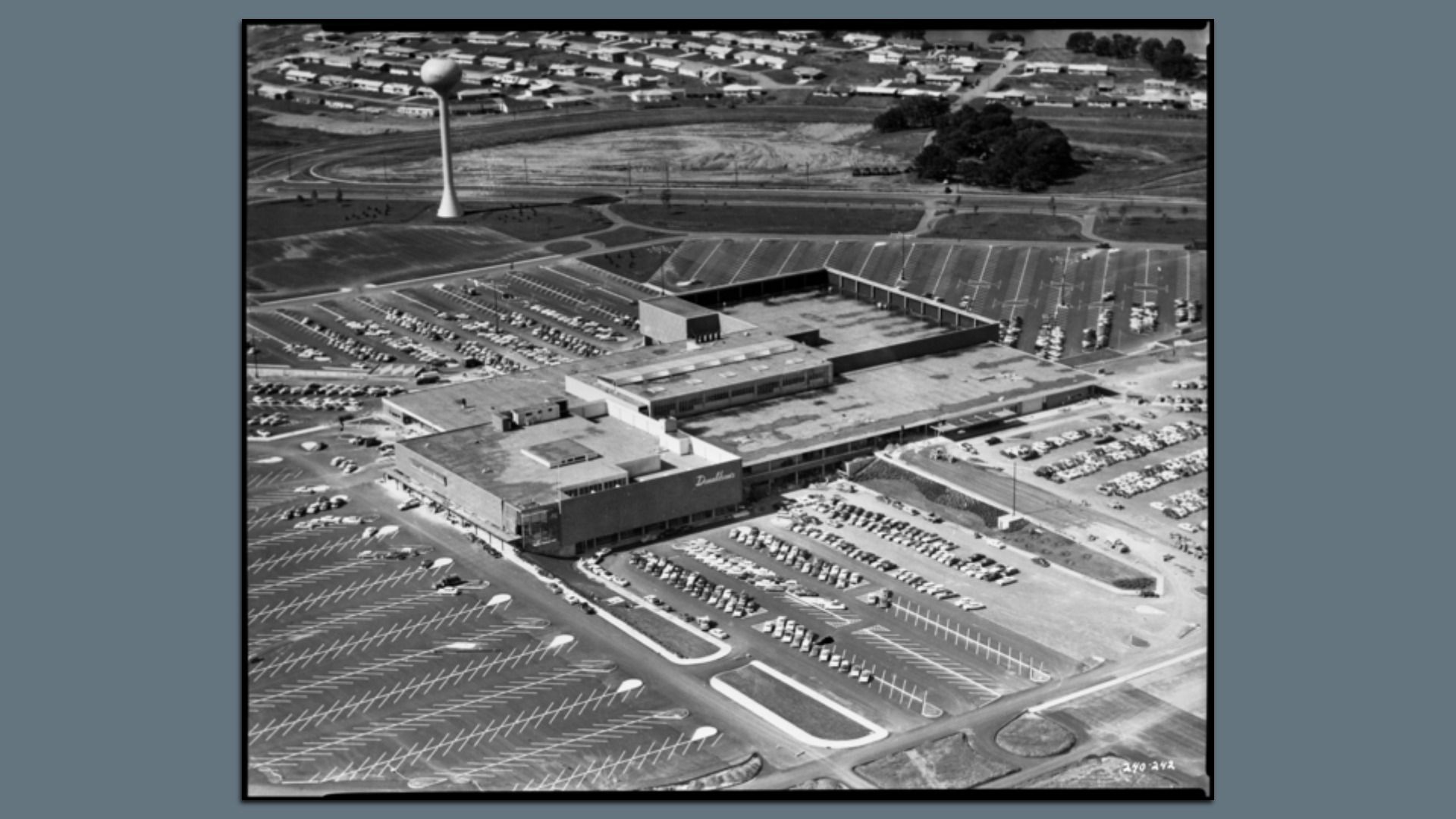 Aerial black-and-white view of a large shopping complex with a long flat-roofed building, crowded parking lots, a tall water tower nearby, and surrounding fields and houses.