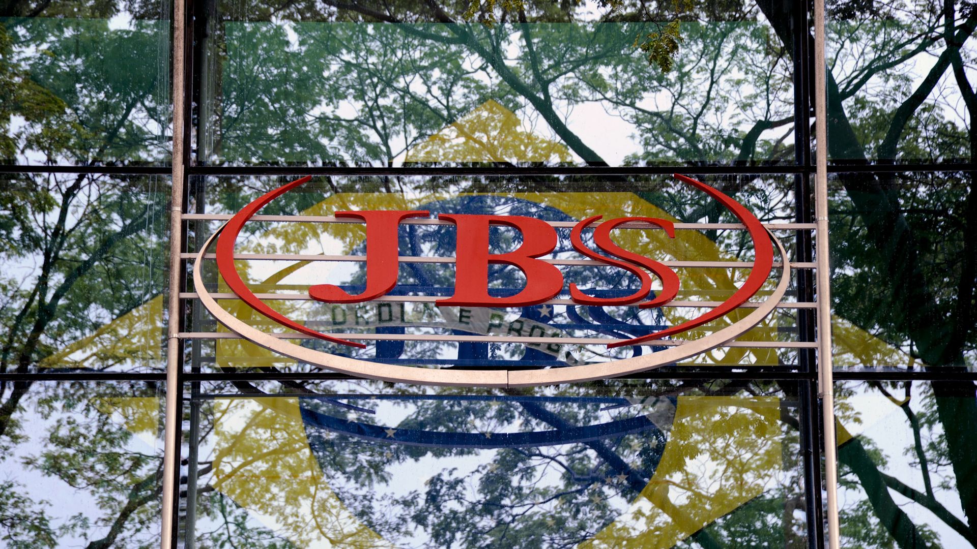 Glass storefront reflecting green trees, with a large red CBS oval logo across the center. Behind the sign are blue and yellow shapes suggesting a flag in the background.
