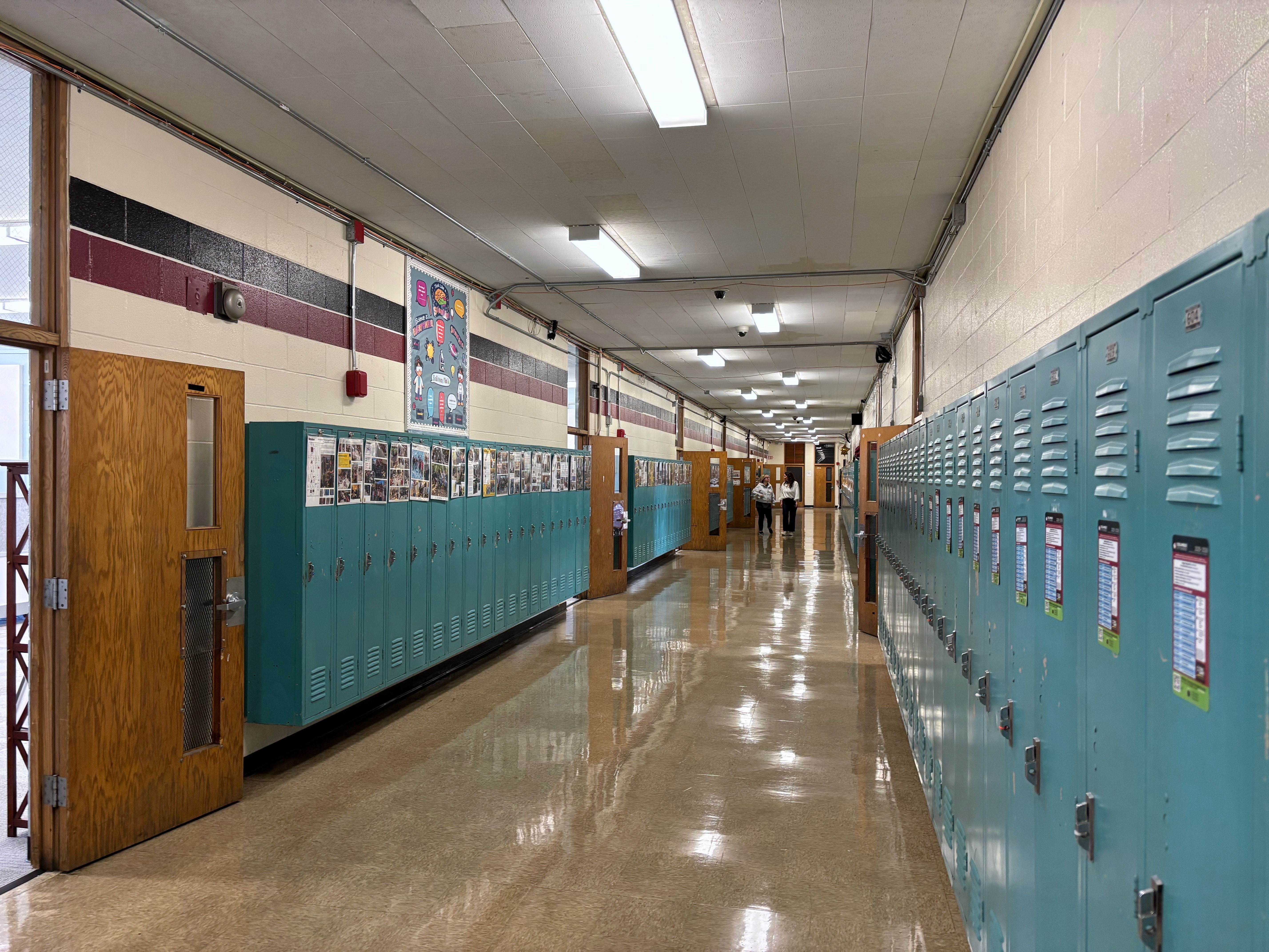 Long school hallway with teal lockers on both sides, a wooden door at left, posters along the walls, and two students talking in the distance under bright fluorescent lights.