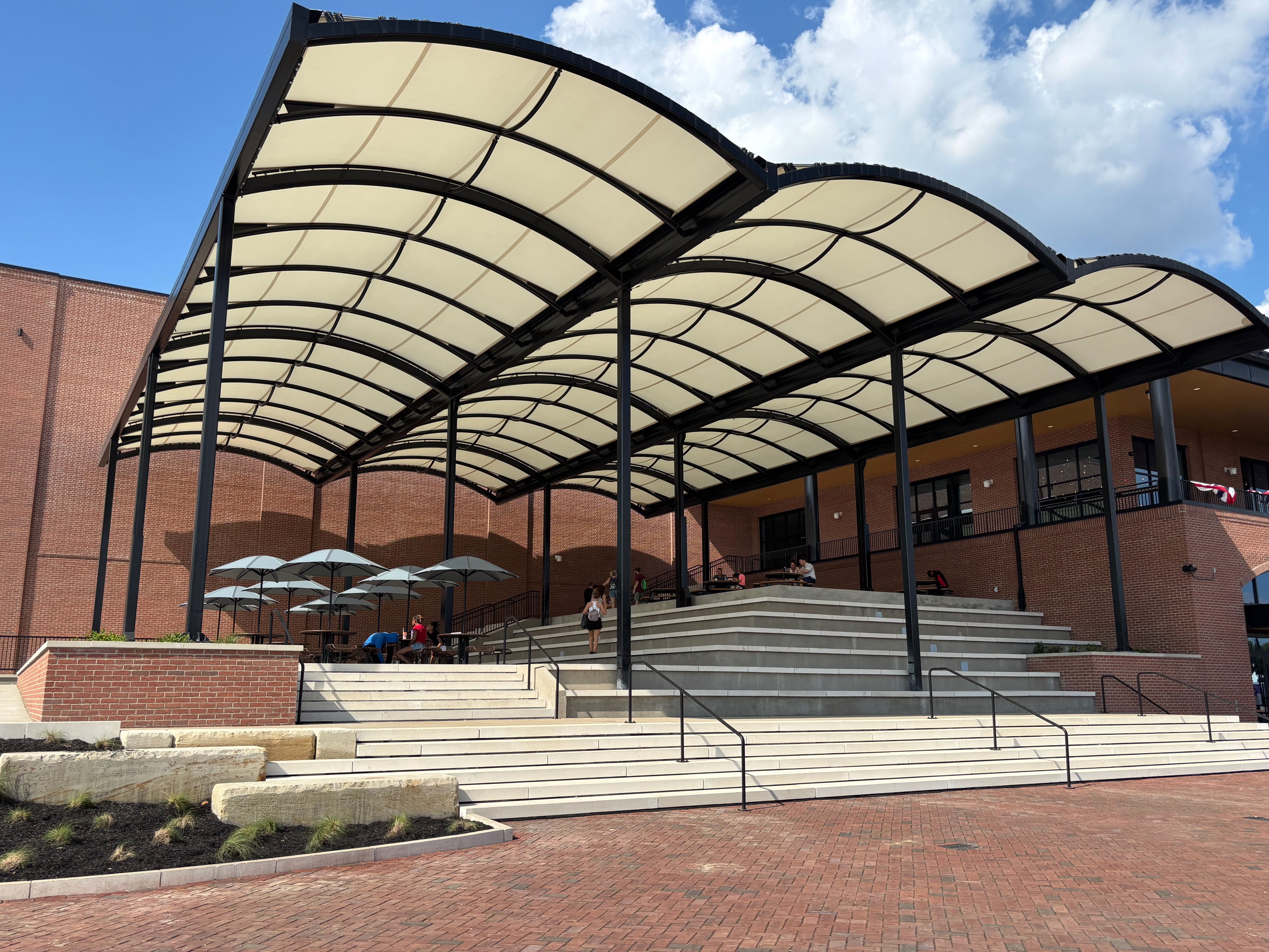 Curved beige translucent canopy with black steel frame arches over a brick building. Wide concrete steps, outdoor seating with umbrellas, and a bright blue sky with fluffy clouds.