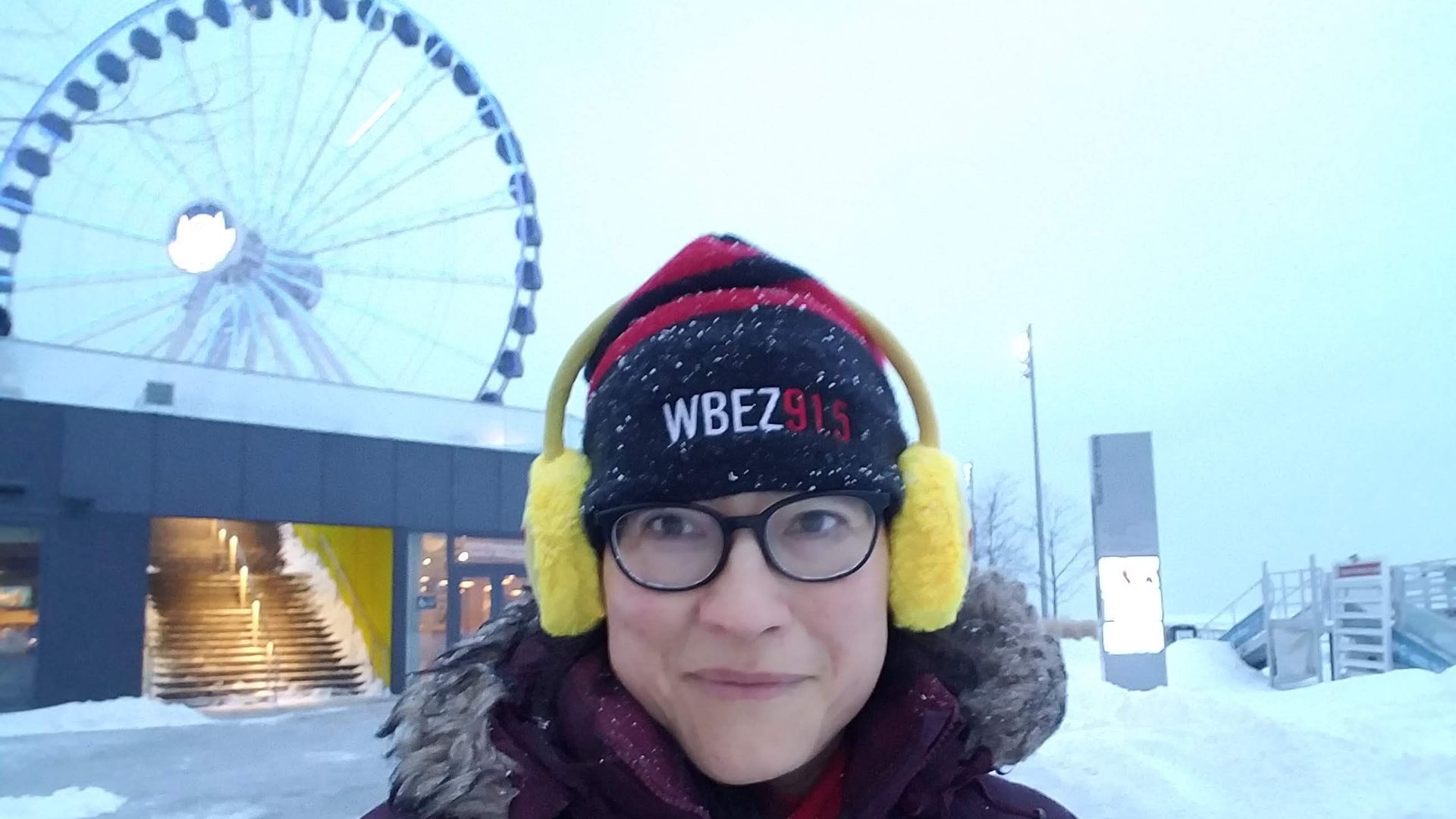 Photo of a woman standing in the snow in front of a ferris wheel. 