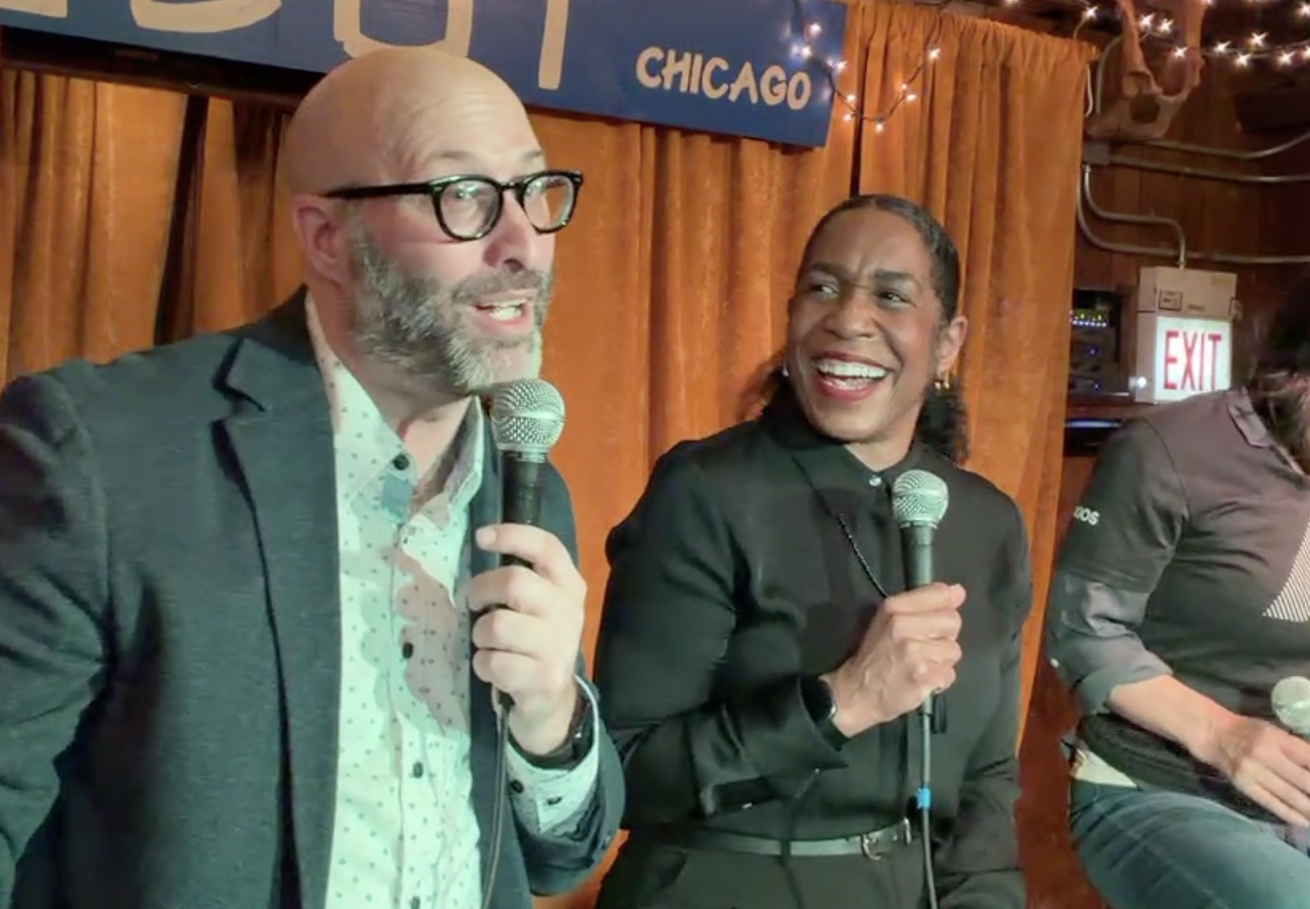 Two speakers on a small stage with orange curtains and string lights. Left: bald man with glasses and a beard in a dark blazer, speaking into a mic. Right: smiling woman in black, also holding a mic.